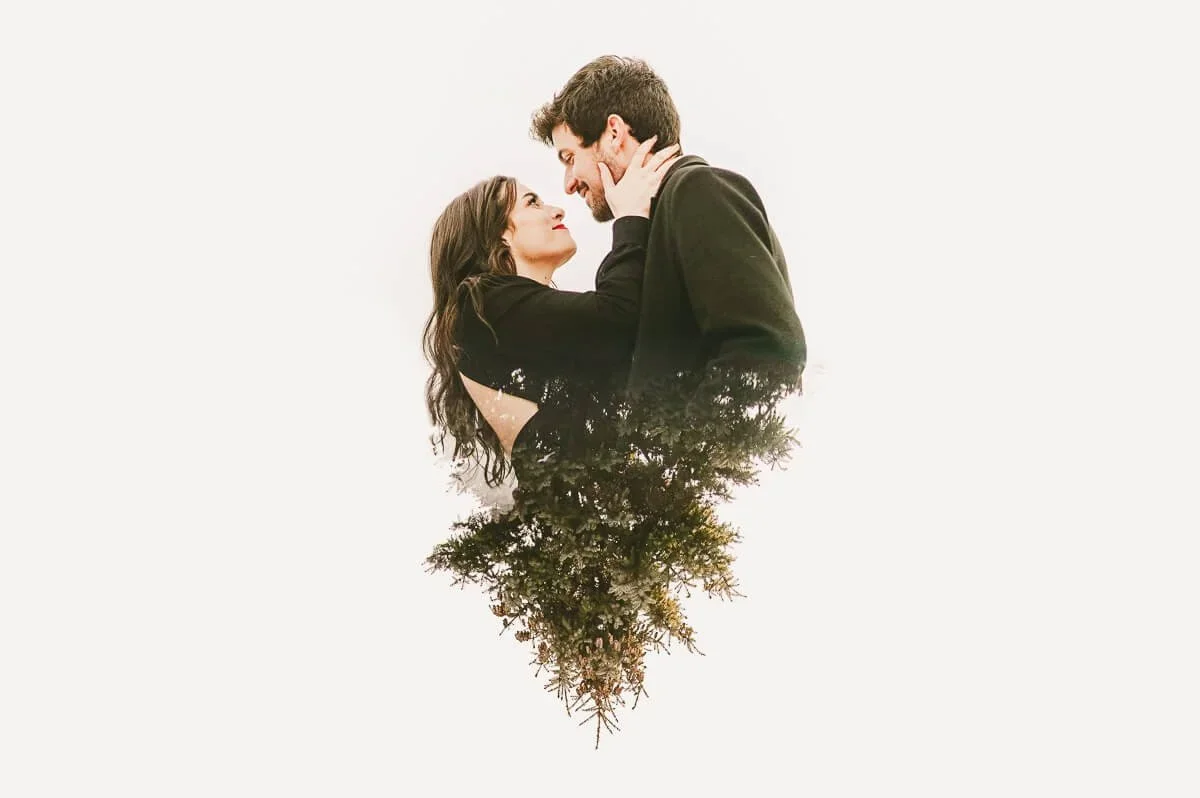 Portrait of a couple wearing black against a white background, smiling at each other, with the lower half of the image featuring a reflection of a tree.