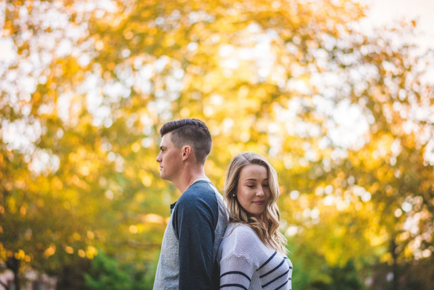 Engaged couple standing back to back during a fall engagement photoshoot surrounded by golden autumn leaves.
