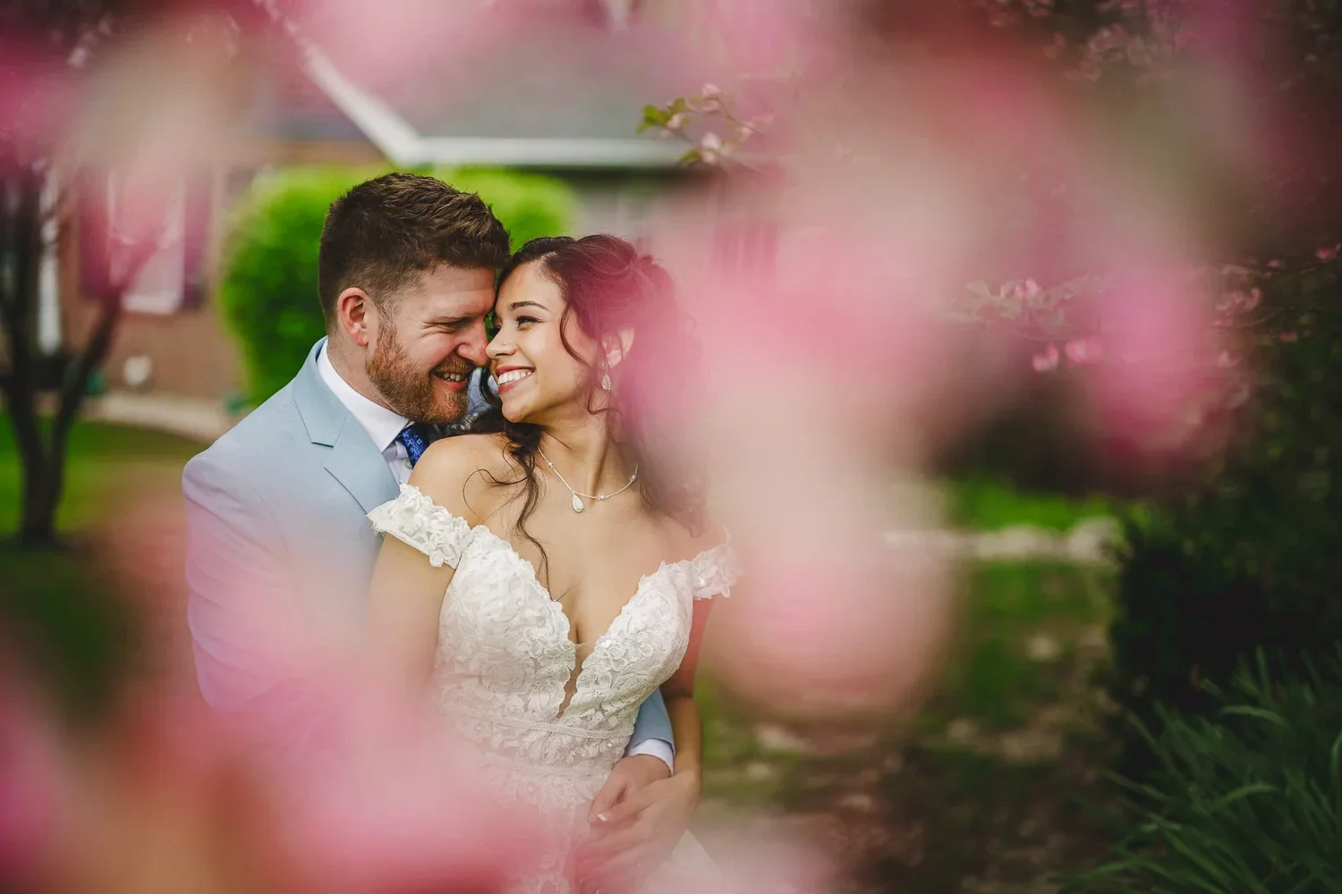 Bride and groom embracing and smiling on their wedding day with soft pink flowers blurred in the foreground.