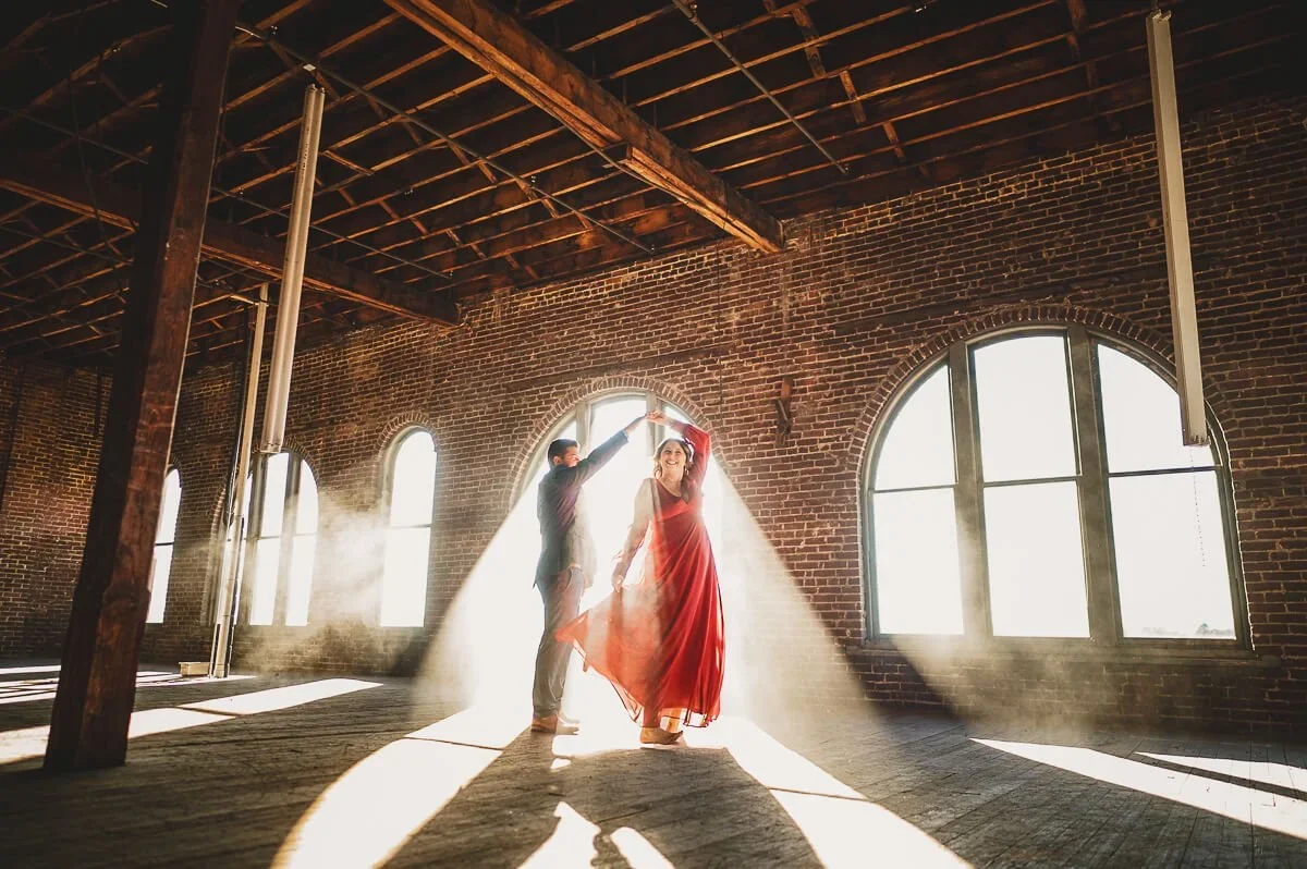 Couple dancing inside an old brick building, with the woman wearing a red dress and twirling as sunlight pours through the windows.