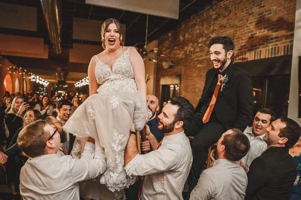 Bride and groom lifted into the air on chairs during their wedding reception, smiling and laughing as guests celebrate.
