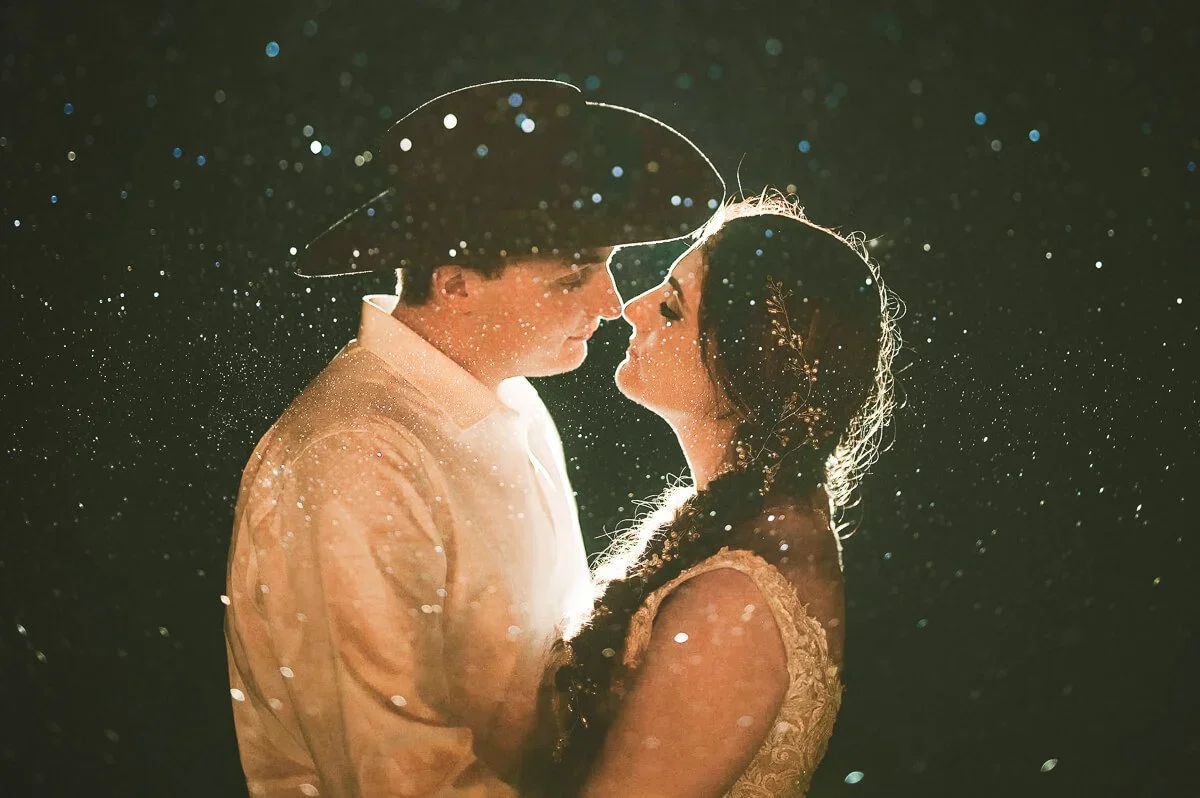 Bride and groom sharing an intimate moment while facing each other, featuring a groom in a black cowboy hat, a bride with braids, and glowing lights around them.