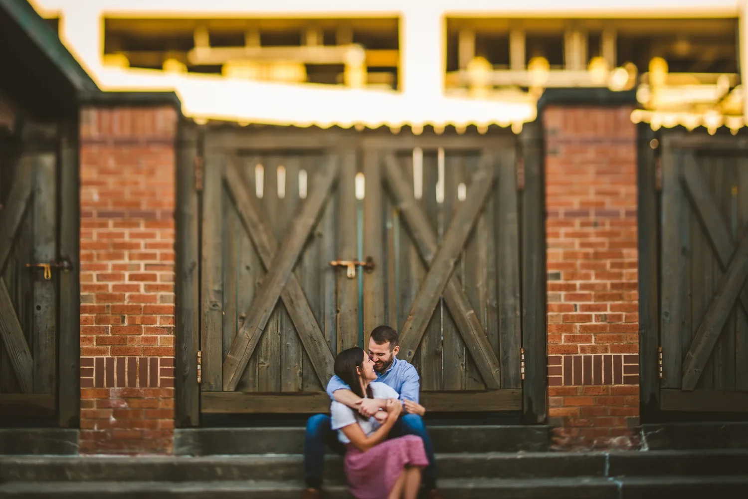 Engagement portrait of a couple sitting on steps outside double doors with a red brick wall behind them.