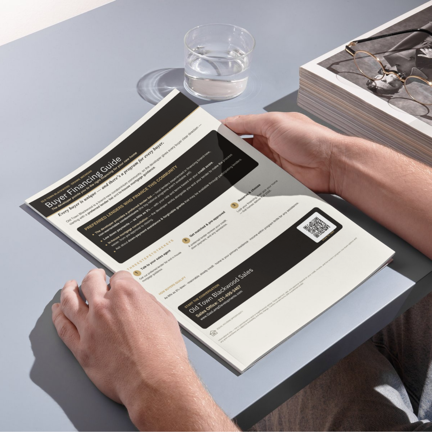 Person holding a printed flyer or brochure about financial guidance sitting at a table next to a glass of water and a stack of magazines or books with eyeglasses on top.