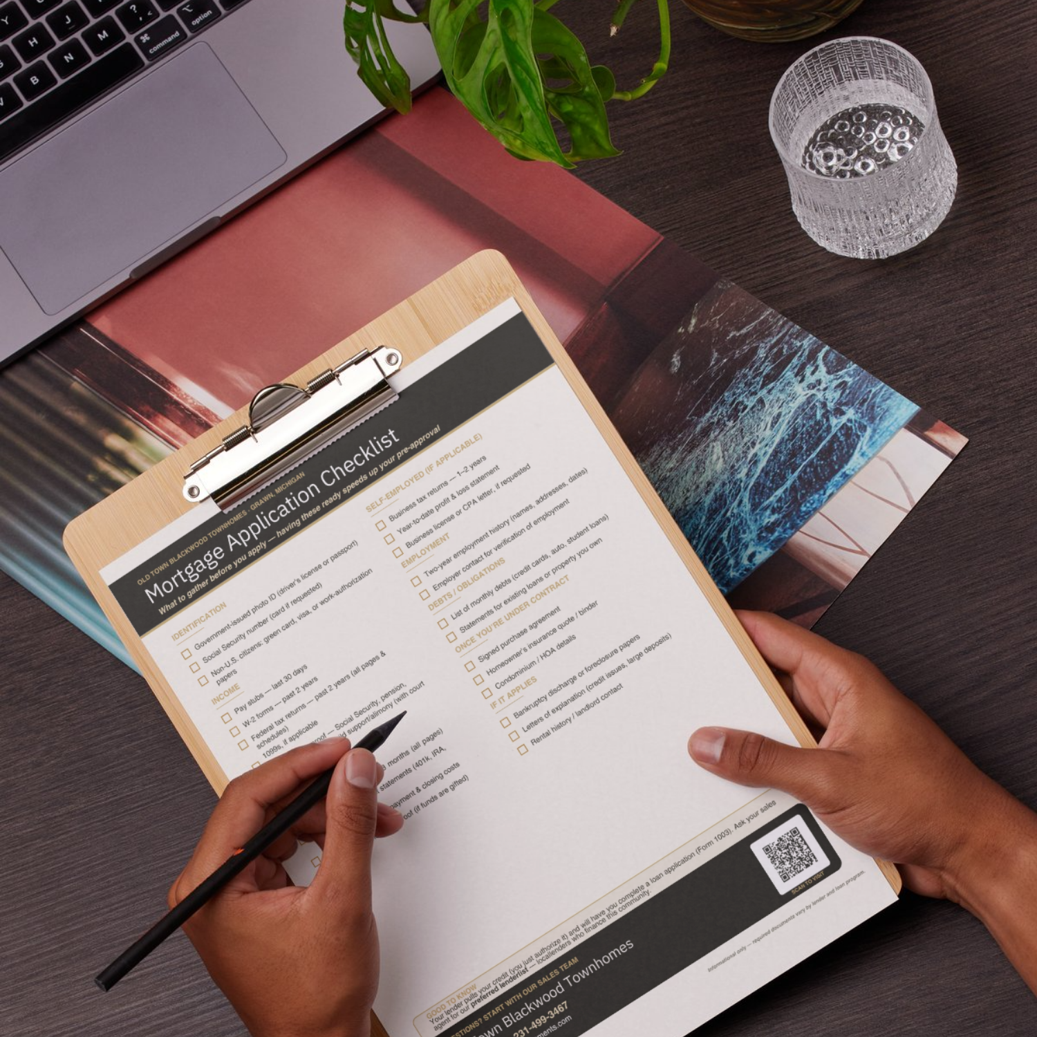 Hands holding a pen and filling out a mortgage application checklist on a clipboard, placed on a wooden desk with a laptop, a glass of water, a plant, and some photographs.