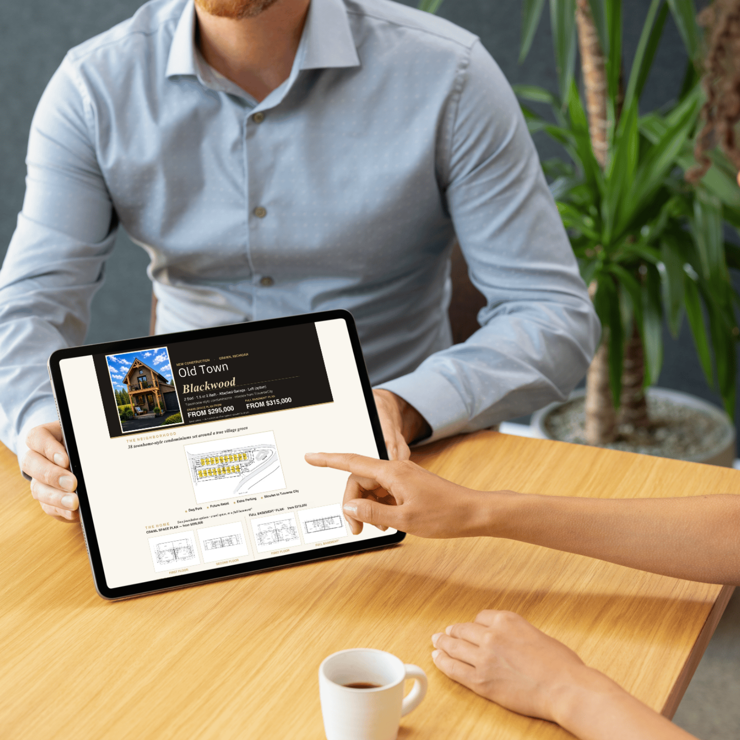 Person showing real estate listings of Old Town Blackwood on a tablet to another person sitting at a wooden table, with a cup of coffee nearby and a plant in the background.