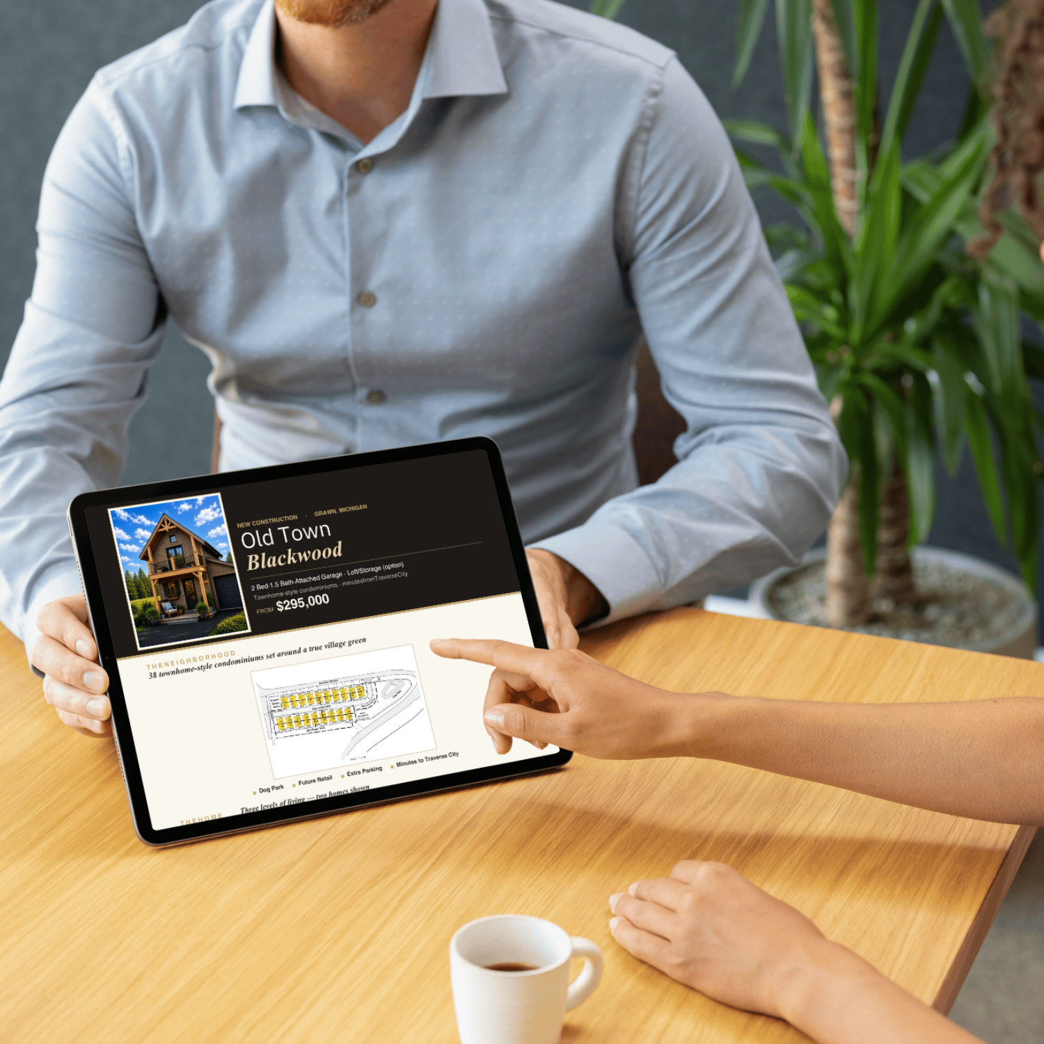 A person in a light gray shirt is showing a tablet screen about real estate listings to another individual with a light-colored hand. The tablet displays a property listing for 'Old Town Blackwood' in Grand Rapids, Michigan, with a picture of a house and details including price and features. There is a wooden table with a white coffee mug in the foreground and a green plant in the background.