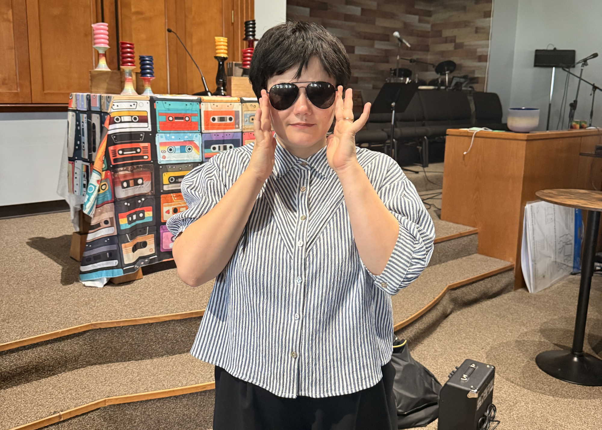 Person with short dark hair wearing sunglasses, a striped button-up shirt, and a wedding ring, adjusting their glasses in a room with musical equipment and colorful cassette tape art in the background.
