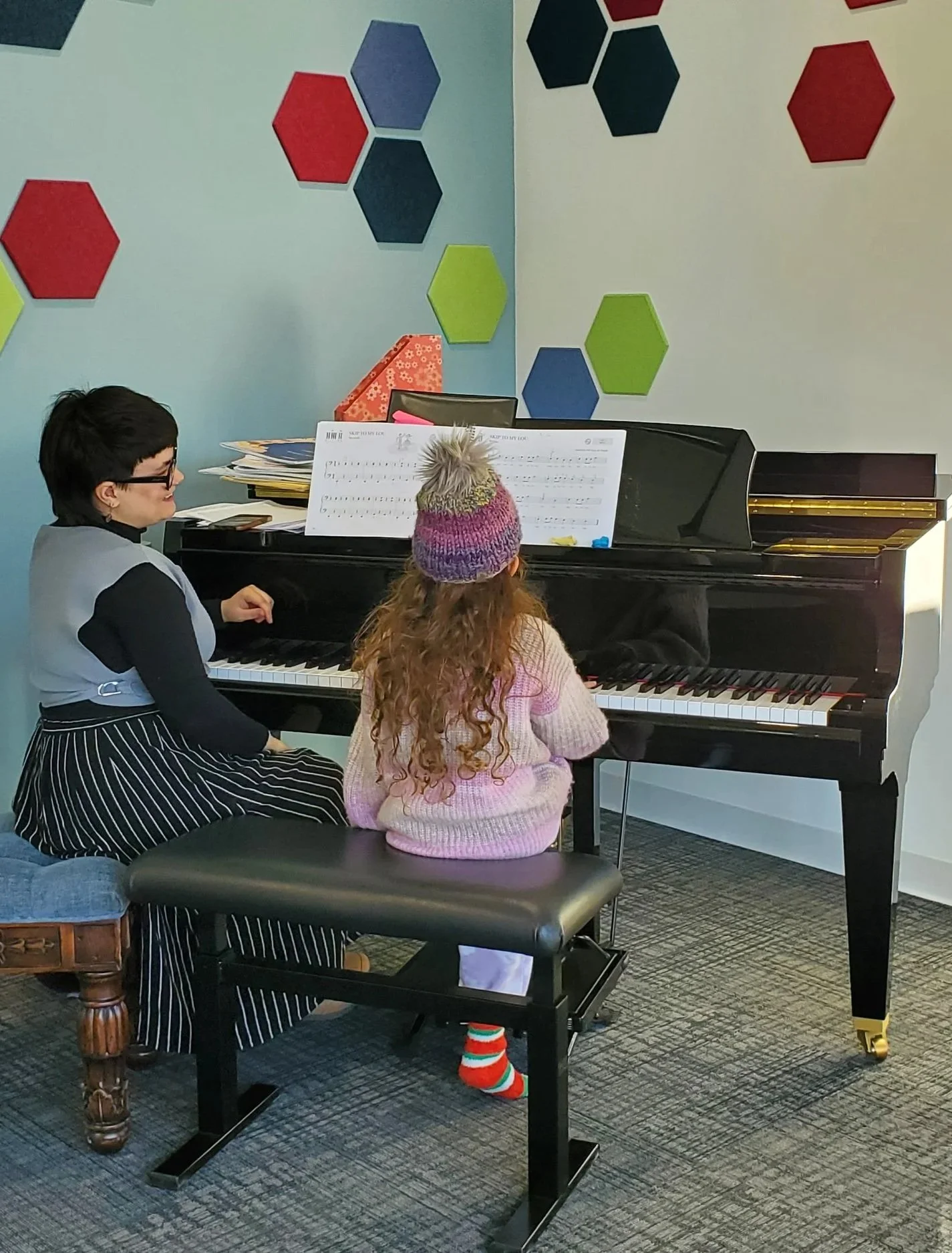 Two children sitting at a black piano; one girl with curly hair wearing a pink sweater and colorful socks, and another girl with glasses and dark hair playing the piano. The room has colorful hexagonal wall decorations.