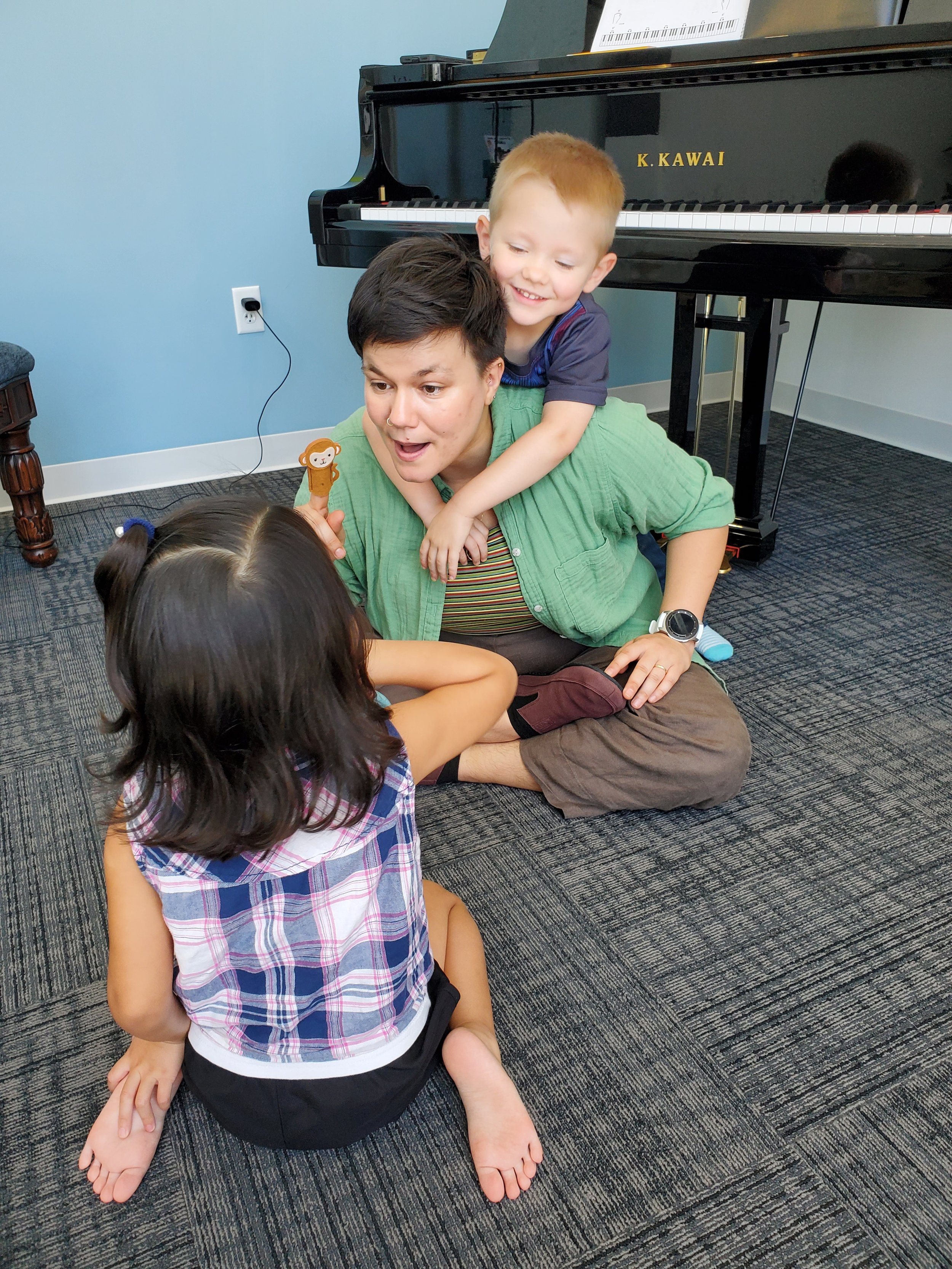 A woman with short dark hair, wearing a green shirt, sitting on the floor, with a young boy on her shoulders, smiling. The boy has short light hair, wearing a dark blue shirt. They are in a room with a black grand piano in the background and a young girl with dark hair in a plaid shirt sitting on the floor in front of them.