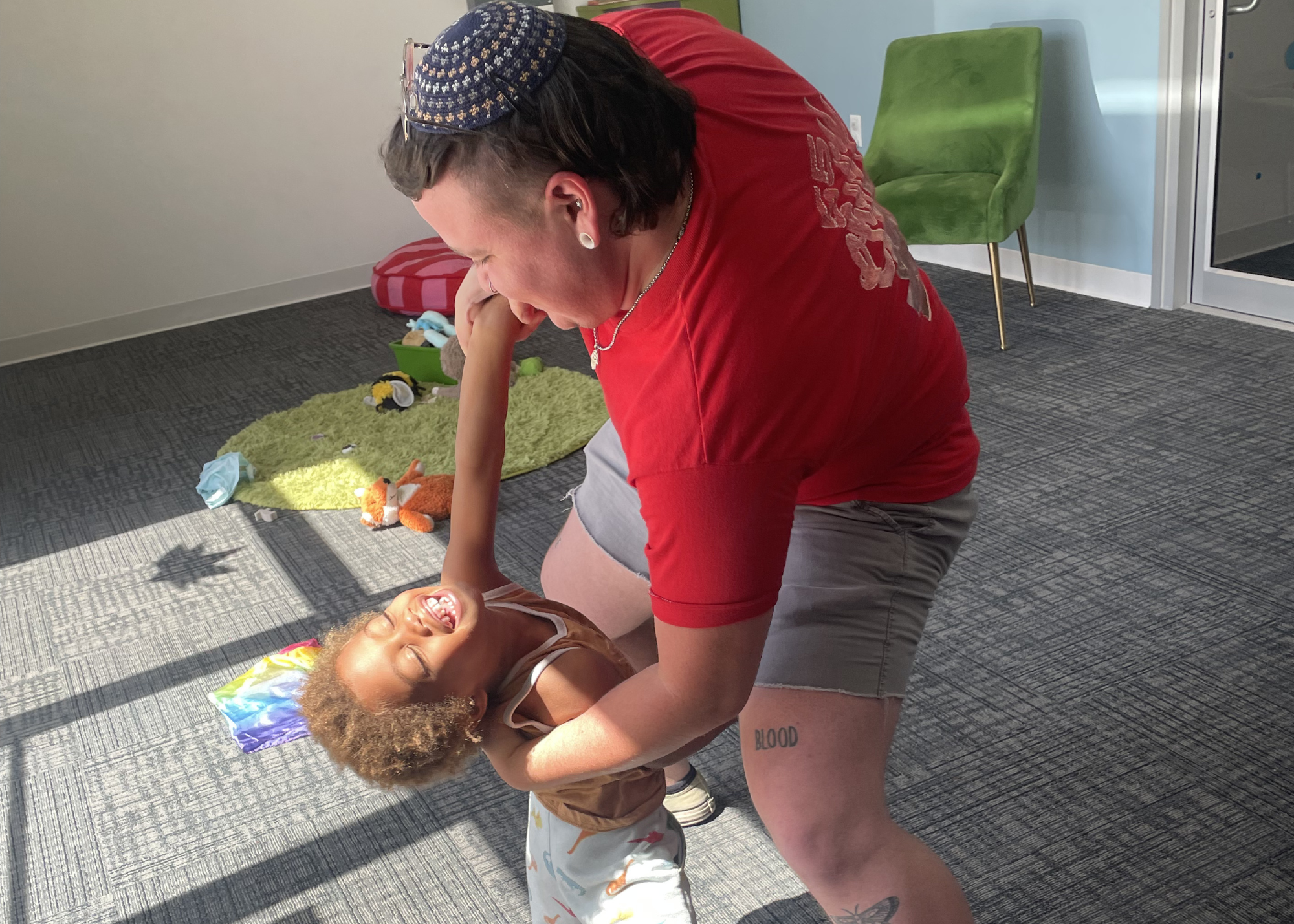 A woman with curly hair lying on the floor, smiling and laughing, wearing a rainbow-colored hat. A man with dark hair, tattoos on his legs, and traditional Jewish clothing, is leaning over her, holding her by the arms and smiling. The setting appears to be an indoor room with toys and a green chair in the background.