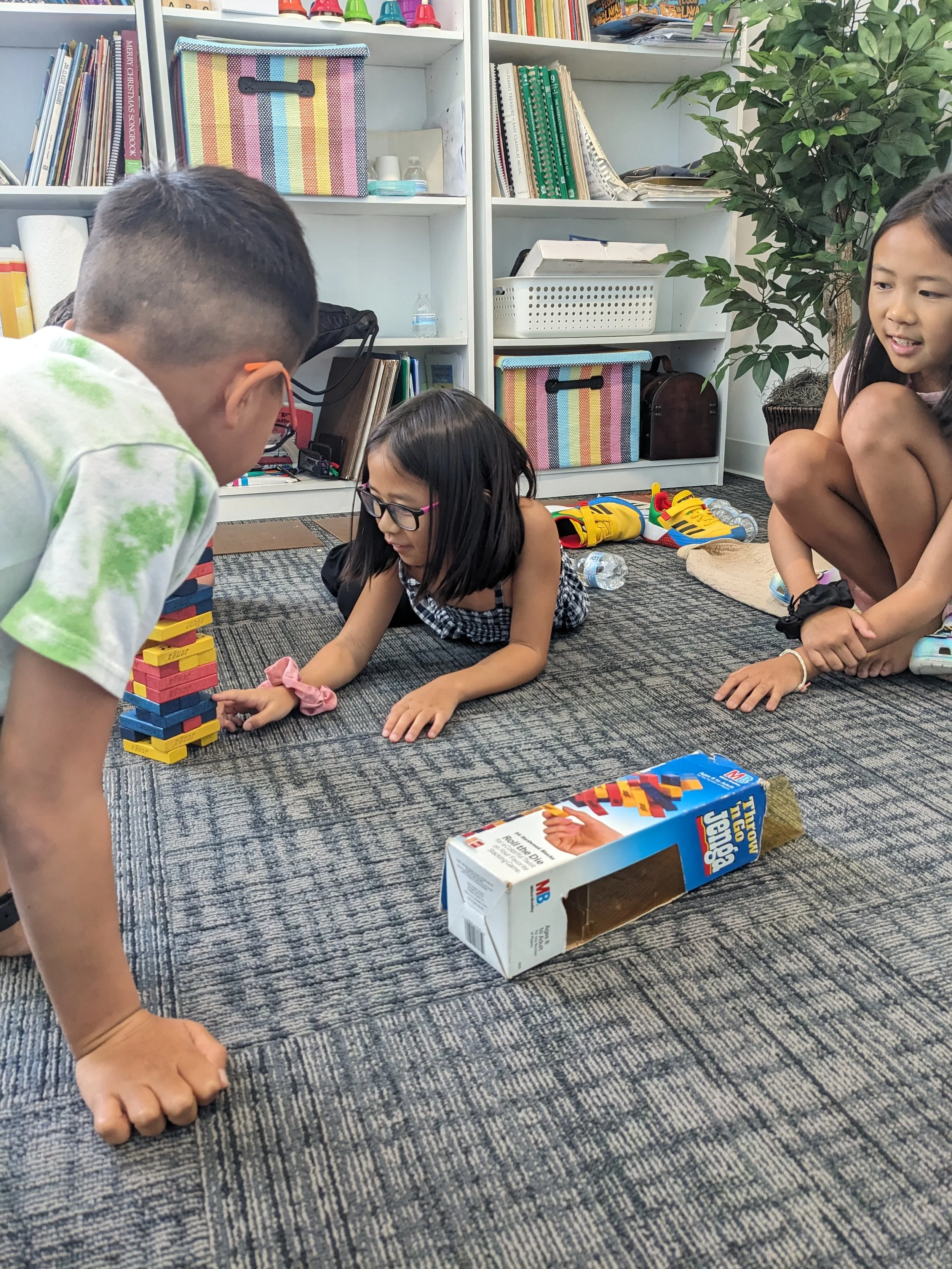 Group of children playing Jenga game on a carpeted floor in a room with bookshelves and green plant in background.