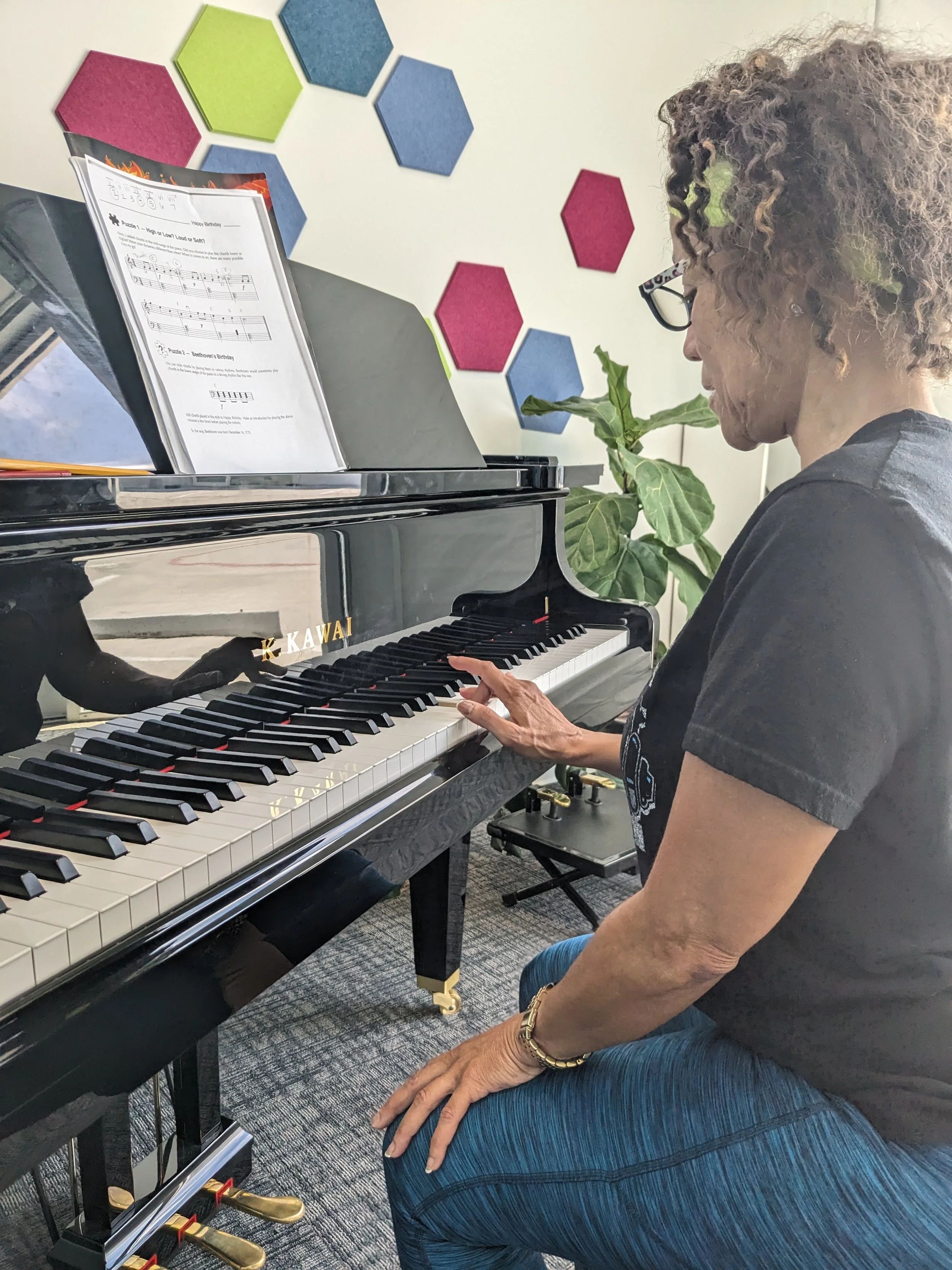 An elderly woman with curly hair and glasses sitting at a black grand piano, playing music composed for a birthday celebration, with a green plant and colorful hexagonal wall decorations in the background.