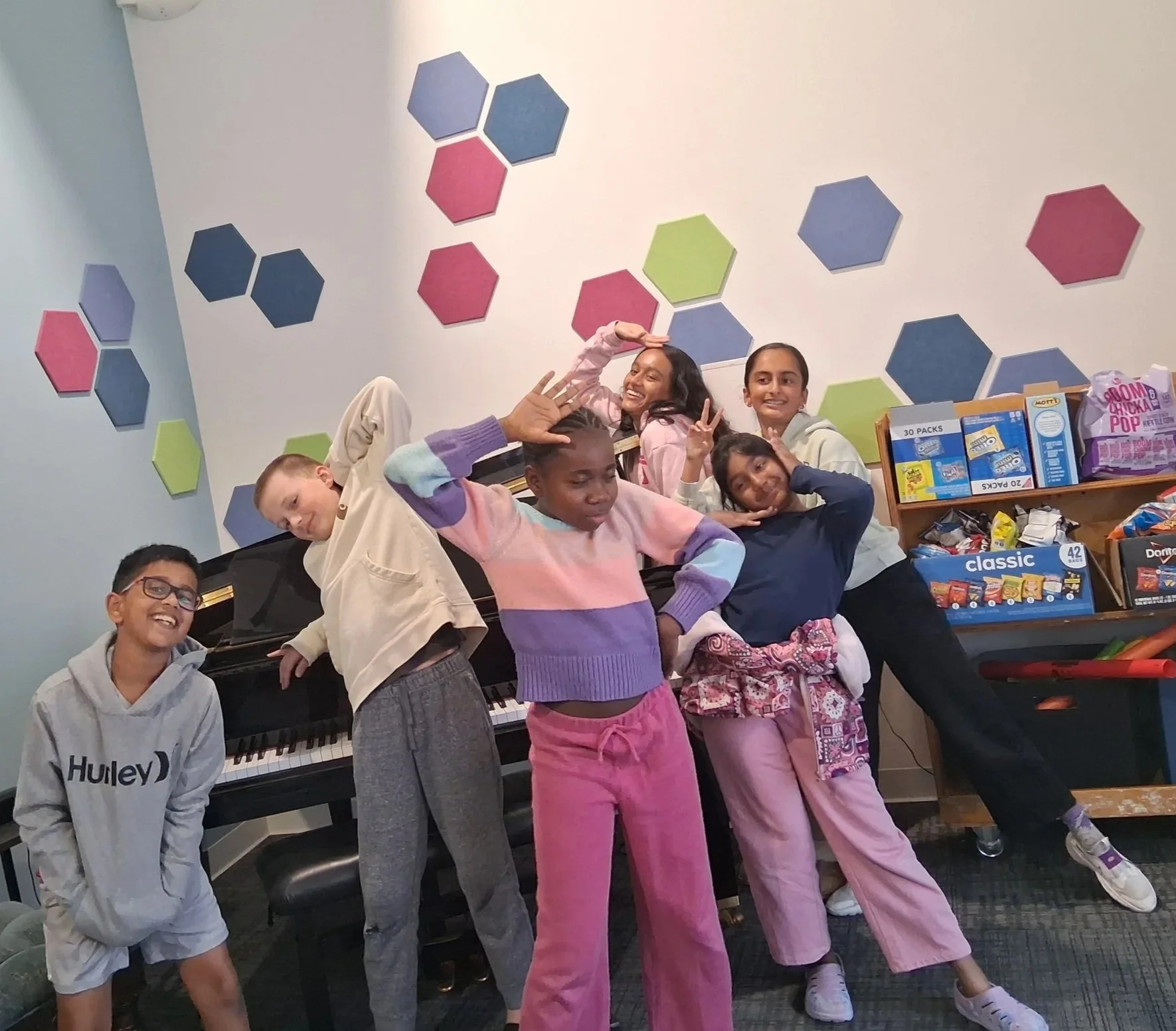 Group of five diverse children posing inside a room with colorful hexagonal wall decorations. Three children are making peace signs and playful gestures. A piano, snack shelves, and a few snacks are visible in the background.