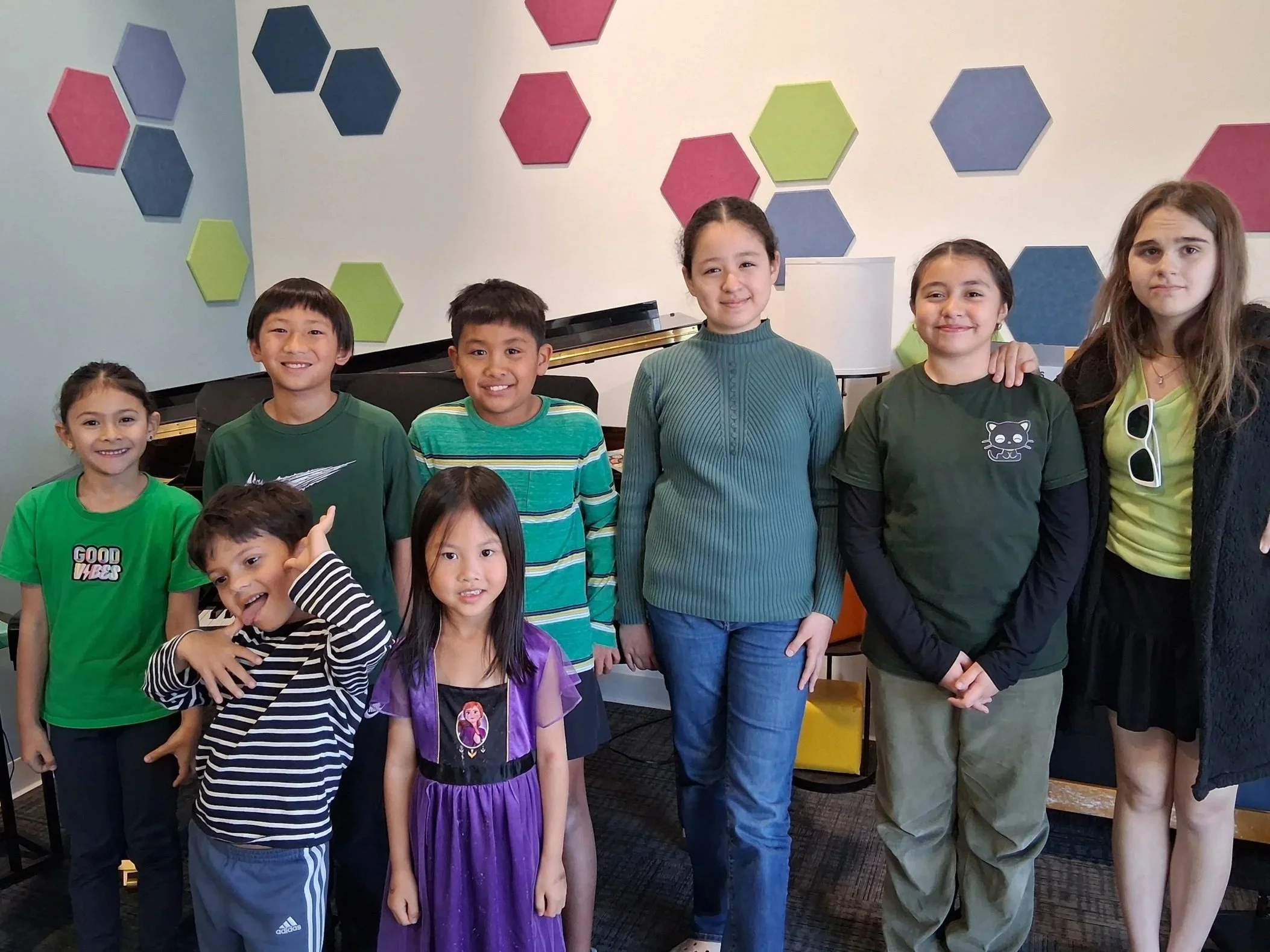 Group of seven children standing together indoors, smiling. They vary in age and ethnic backgrounds. The background features colorful hexagonal wall decorations.