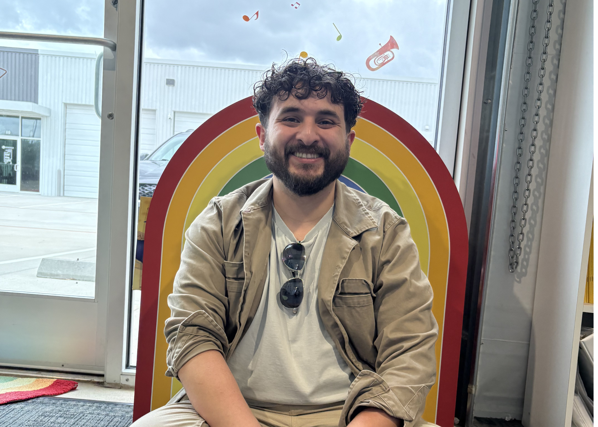 A smiling man with curly hair and a beard sitting in front of a rainbow-shaped decoration with cartoon musical notes on a window. He is wearing a beige jacket, a white t-shirt, and sunglasses hanging from his shirt.