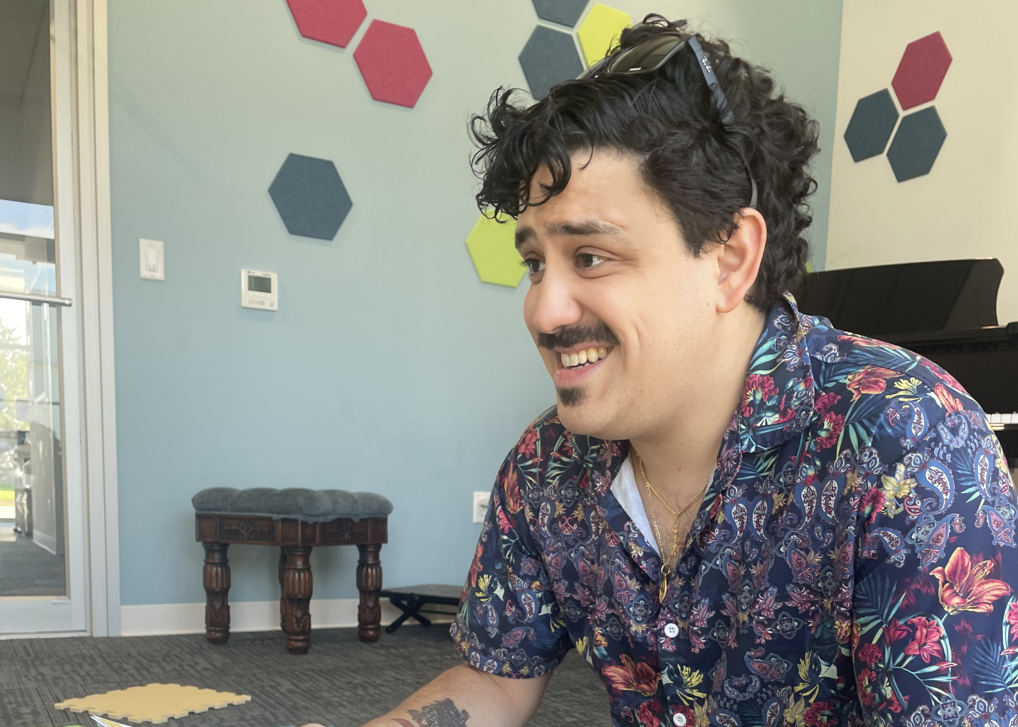 A man with curly black hair, a mustache, and a goatee, smiling, wearing a colorful floral shirt with sunglasses resting on his head, sitting in an indoor space with decorative hexagons on the wall.