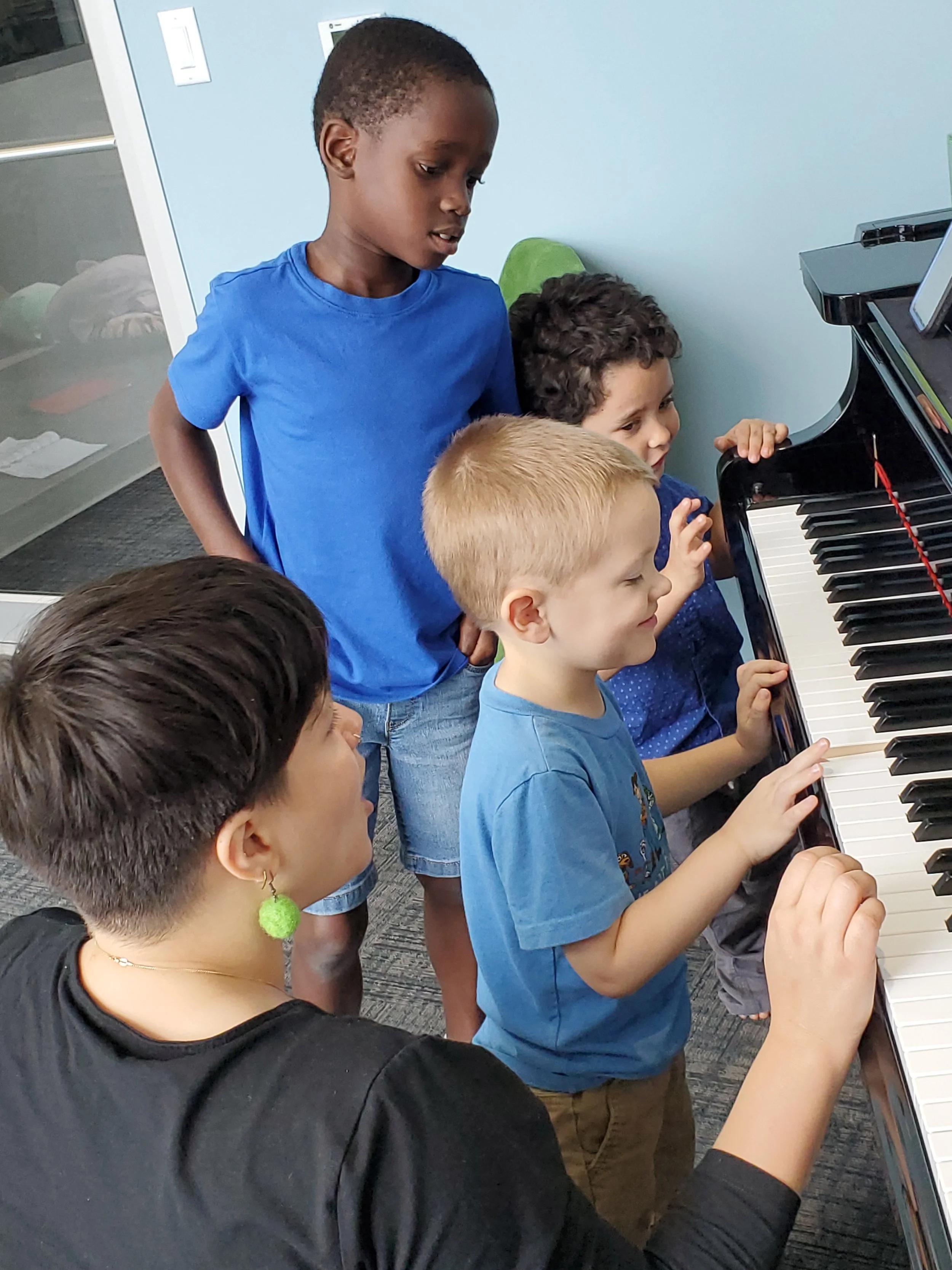 Group of children gathered around a piano, with a woman guiding them. The children are focused on playing or watching, and are in a bright room with light blue walls.