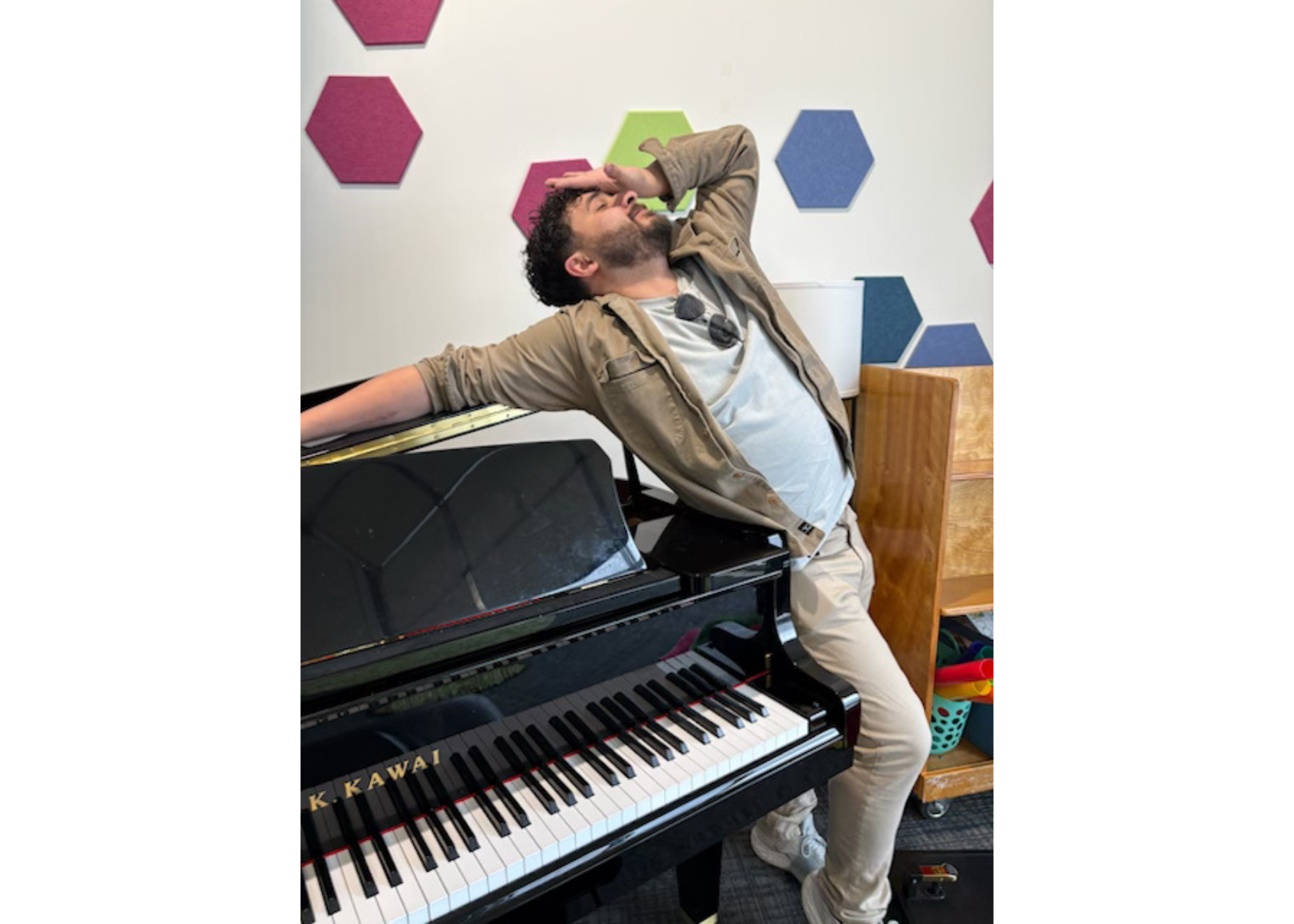A man leaning back with one arm on a piano and the other hand on his forehead, appearing relaxed or exhausted, in a room with colorful hexagonal wall decorations.