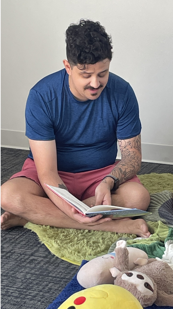 A man with curly dark hair, a mustache, and tattoos on his arms is sitting cross-legged on the floor, reading a book. He is wearing a blue T-shirt and red shorts. There are stuffed toys and a yellow pillow with a smiley face on the floor nearby.