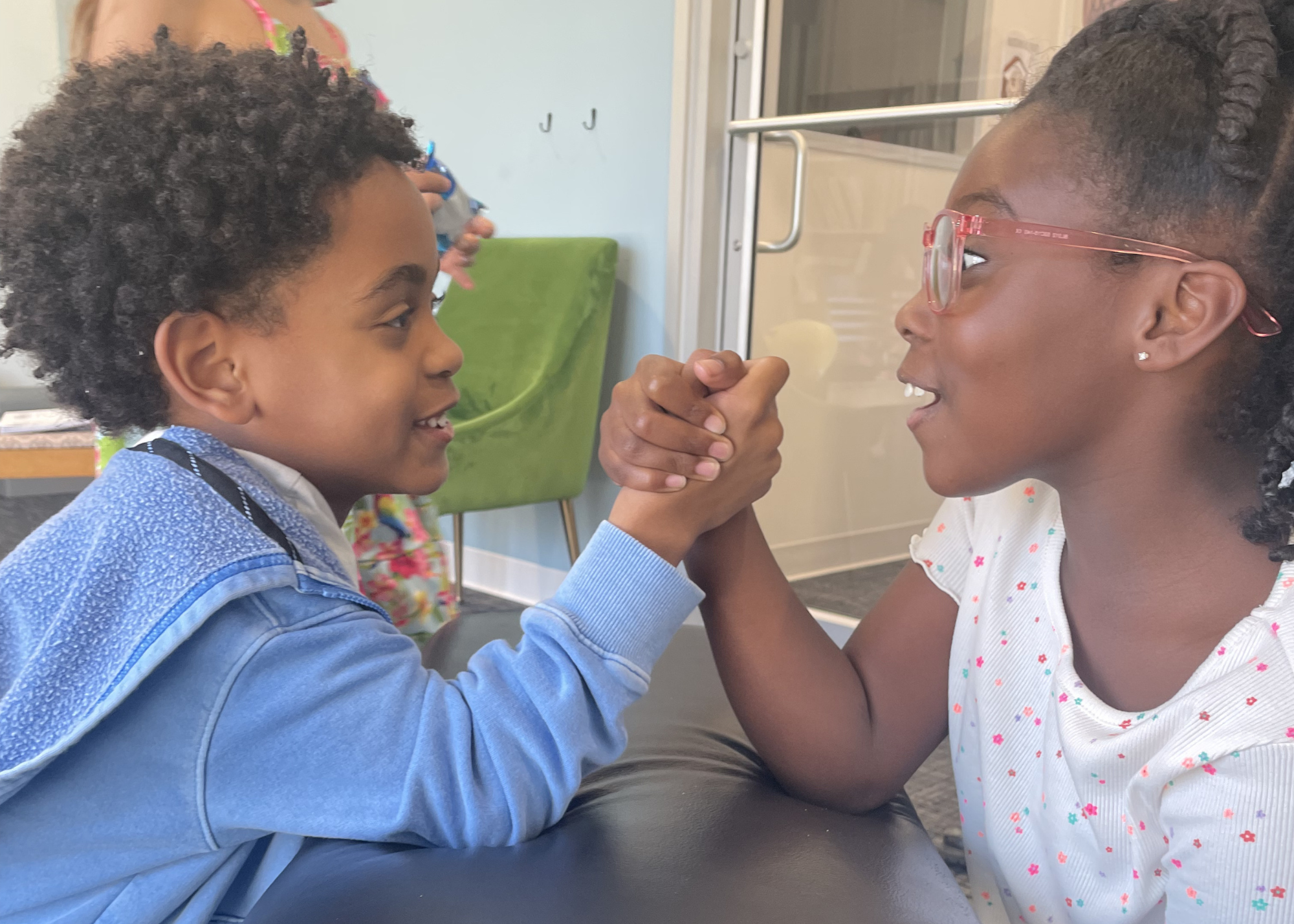 Two children, a boy and a girl, face each other with engaged expressions, holding hands in an arm-wrestling match, inside a room with a green chair and hooks on the wall in the background.