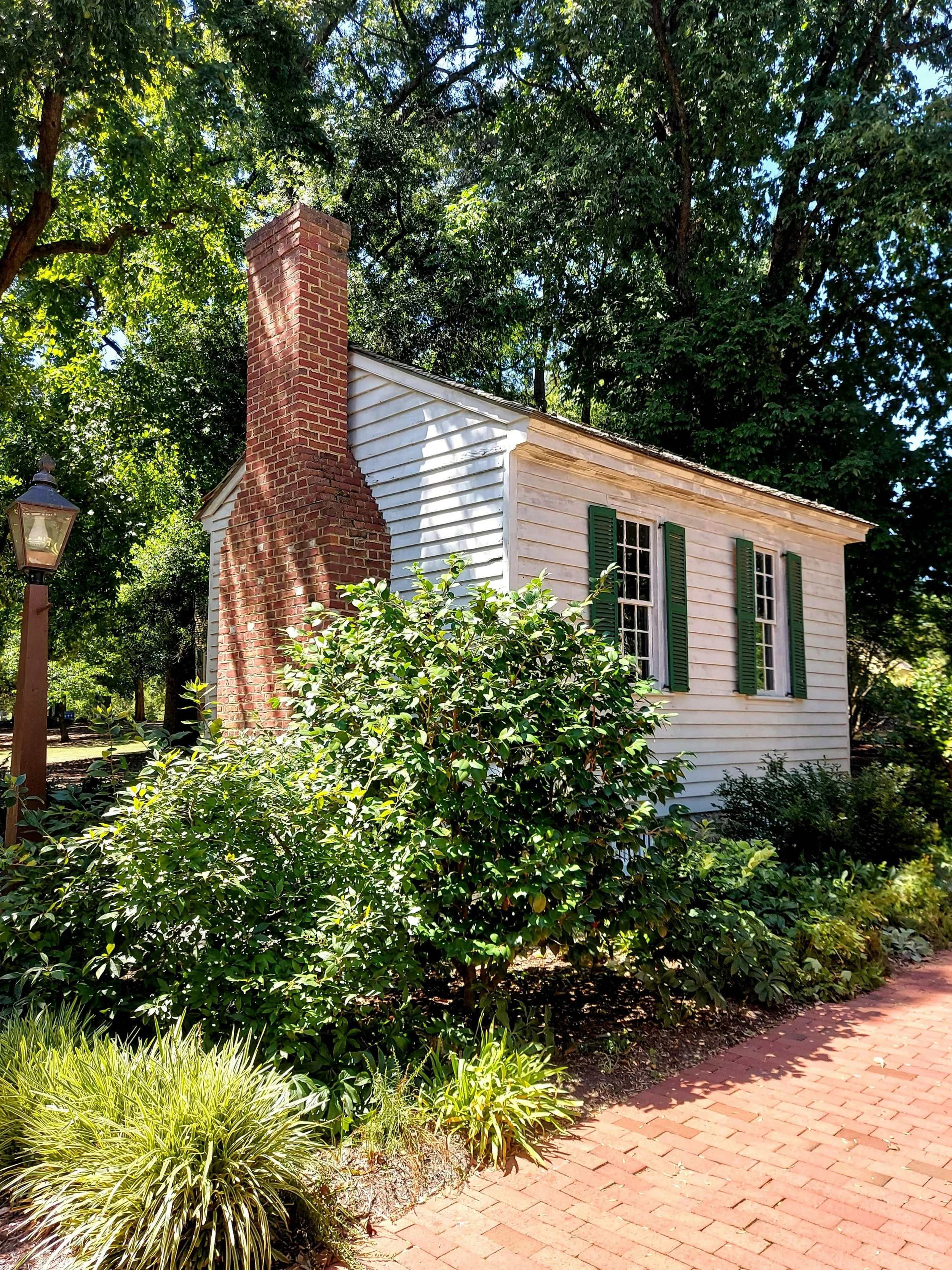 Small house with white wooden siding, three windows with green shutters, and a brick chimney, surrounded by lush green bushes and trees.