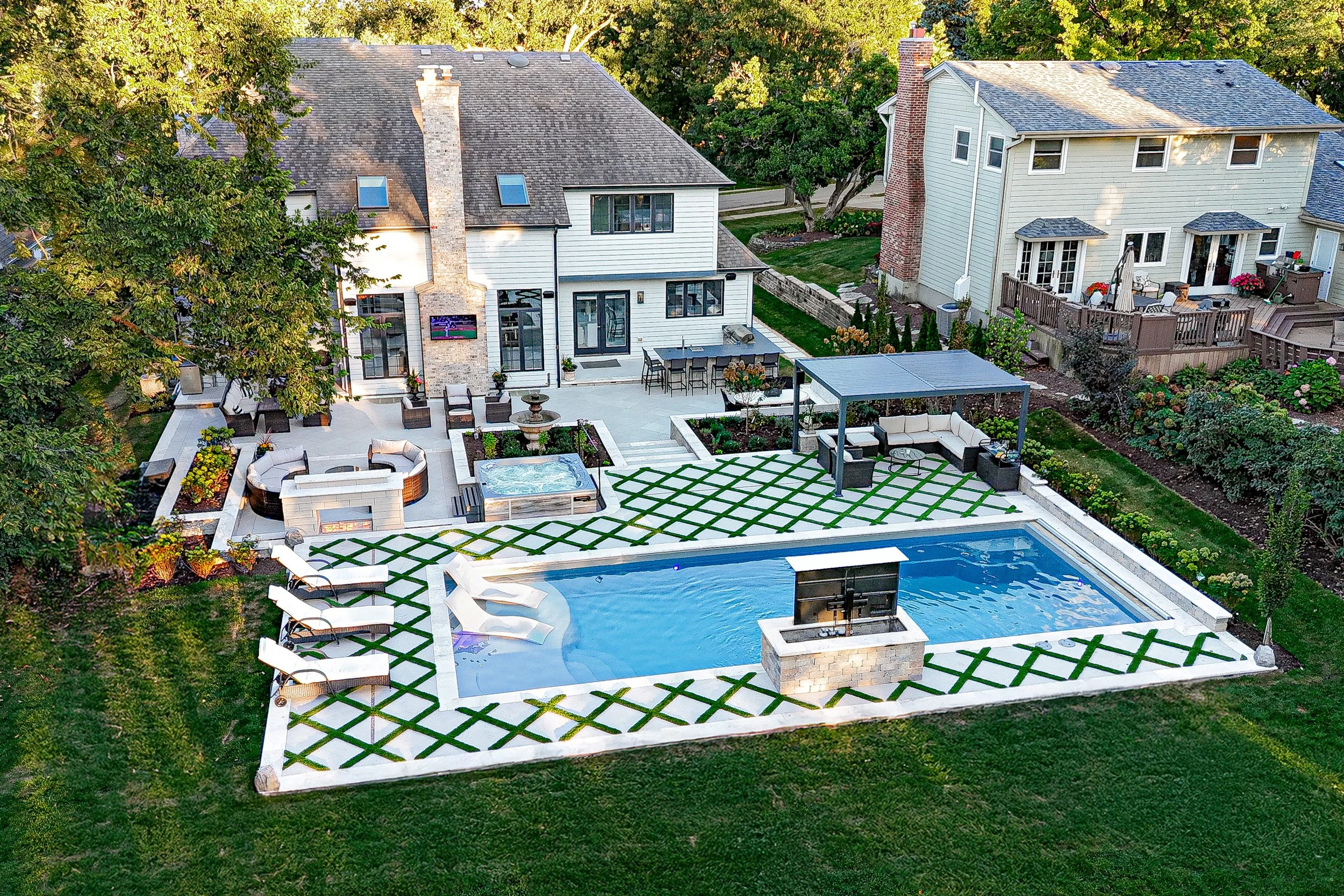 A spacious backyard with a swimming pool, outdoor seating areas including a covered lounge, fire pit, and a hot tub, surrounded by a well-maintained lawn and gardens, adjacent to two houses in a suburban neighborhood.