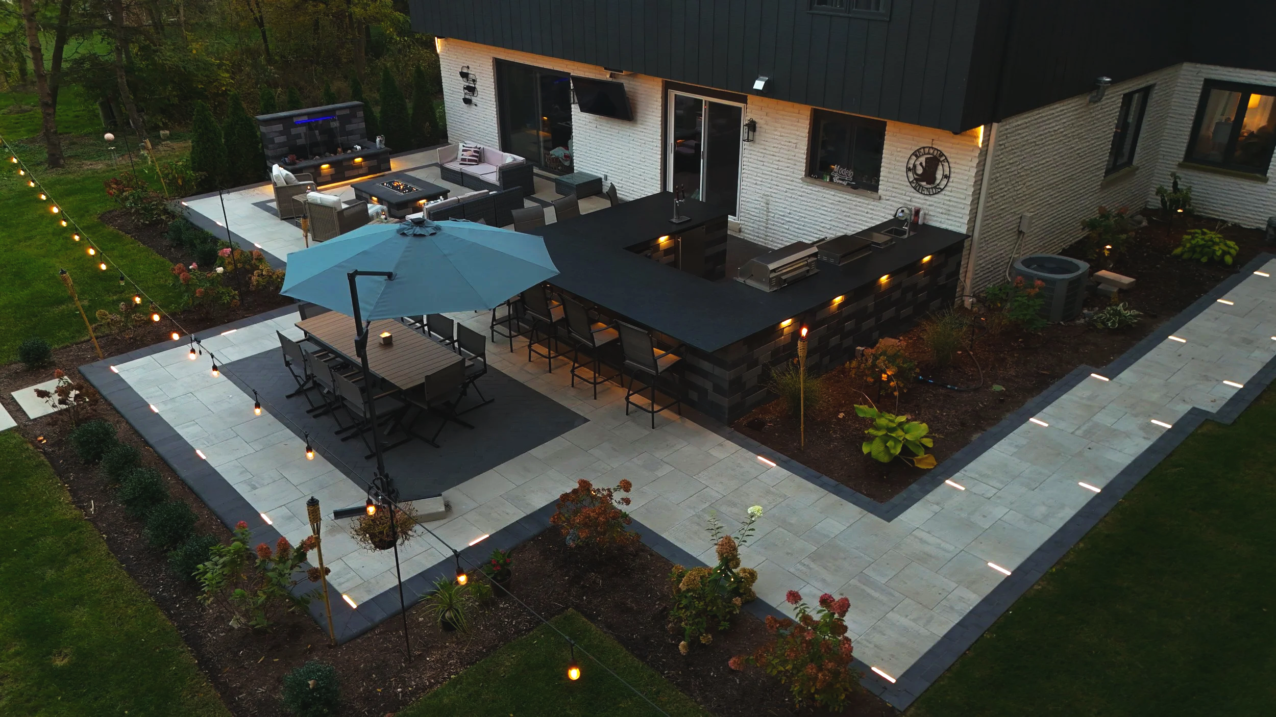 A backyard patio with outdoor furniture, a barbecue area, string lights, an umbrella, and landscaped garden beds at dusk.