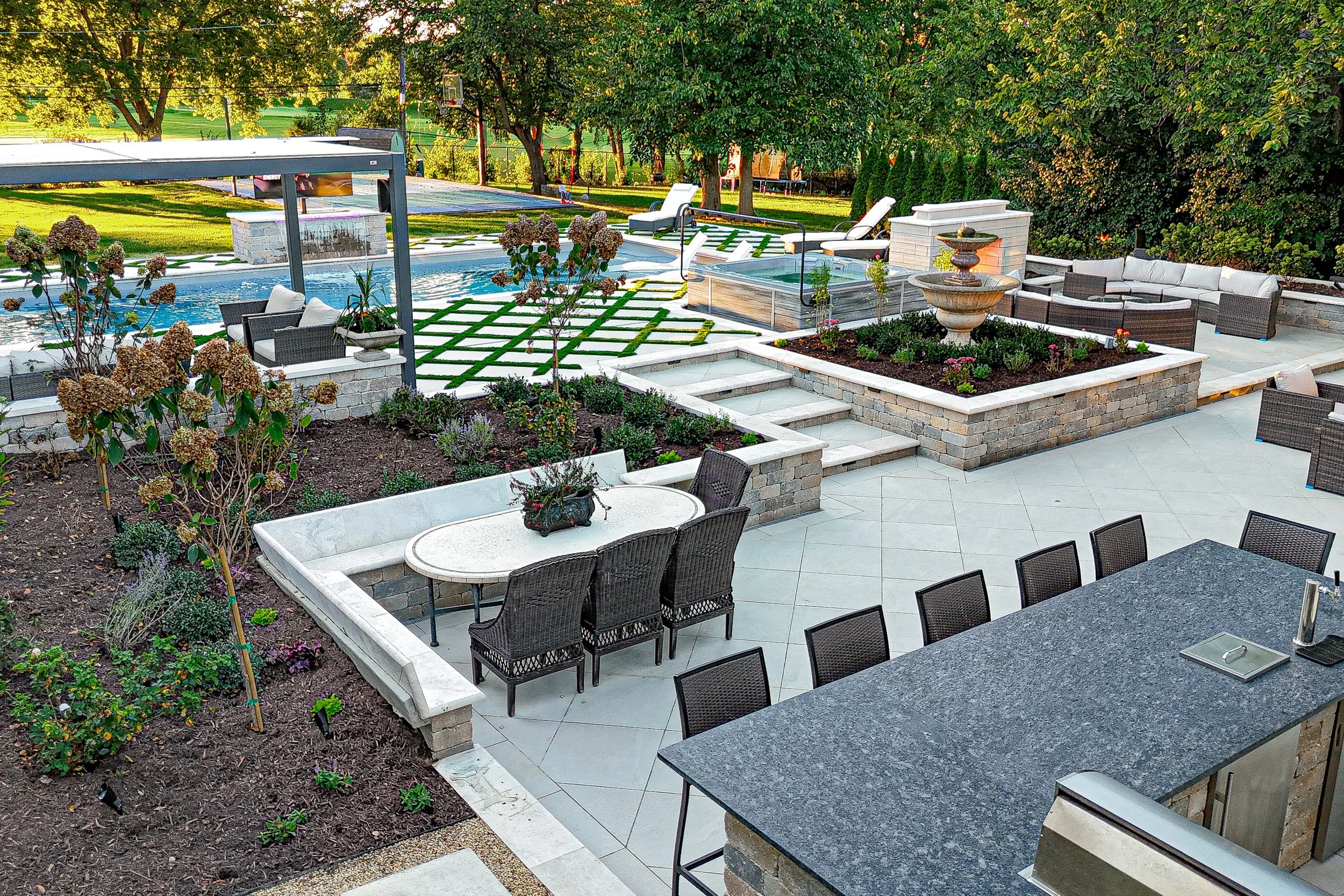 Beautiful backyard with a swimming pool, outdoor seating, garden beds with flowers, and a hot tub surrounded by lush green trees.