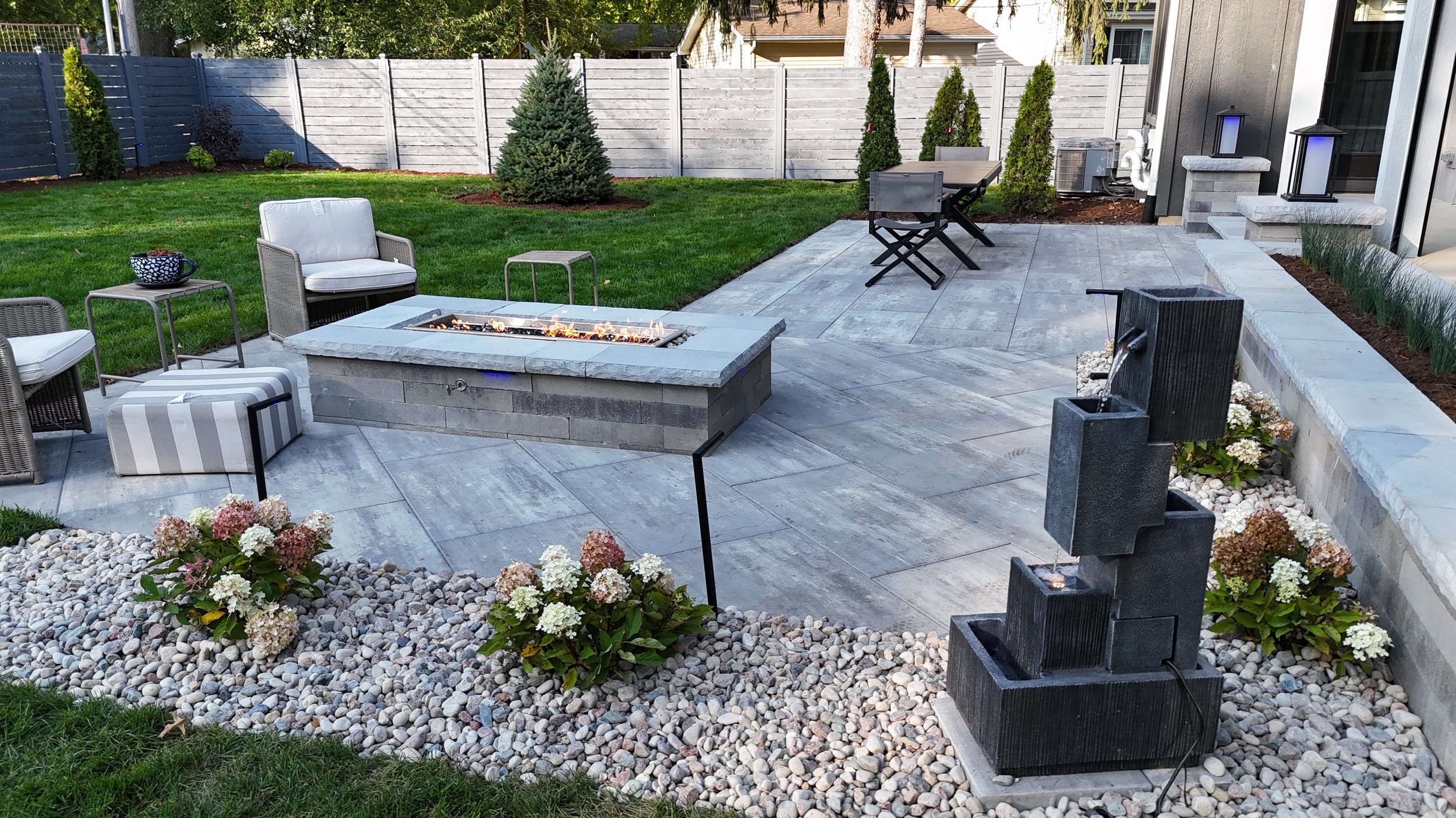 A backyard patio area with outdoor furniture, a fire pit, a water fountain, and landscaped garden beds with flowers and greenery, enclosed by a wooden fence.
