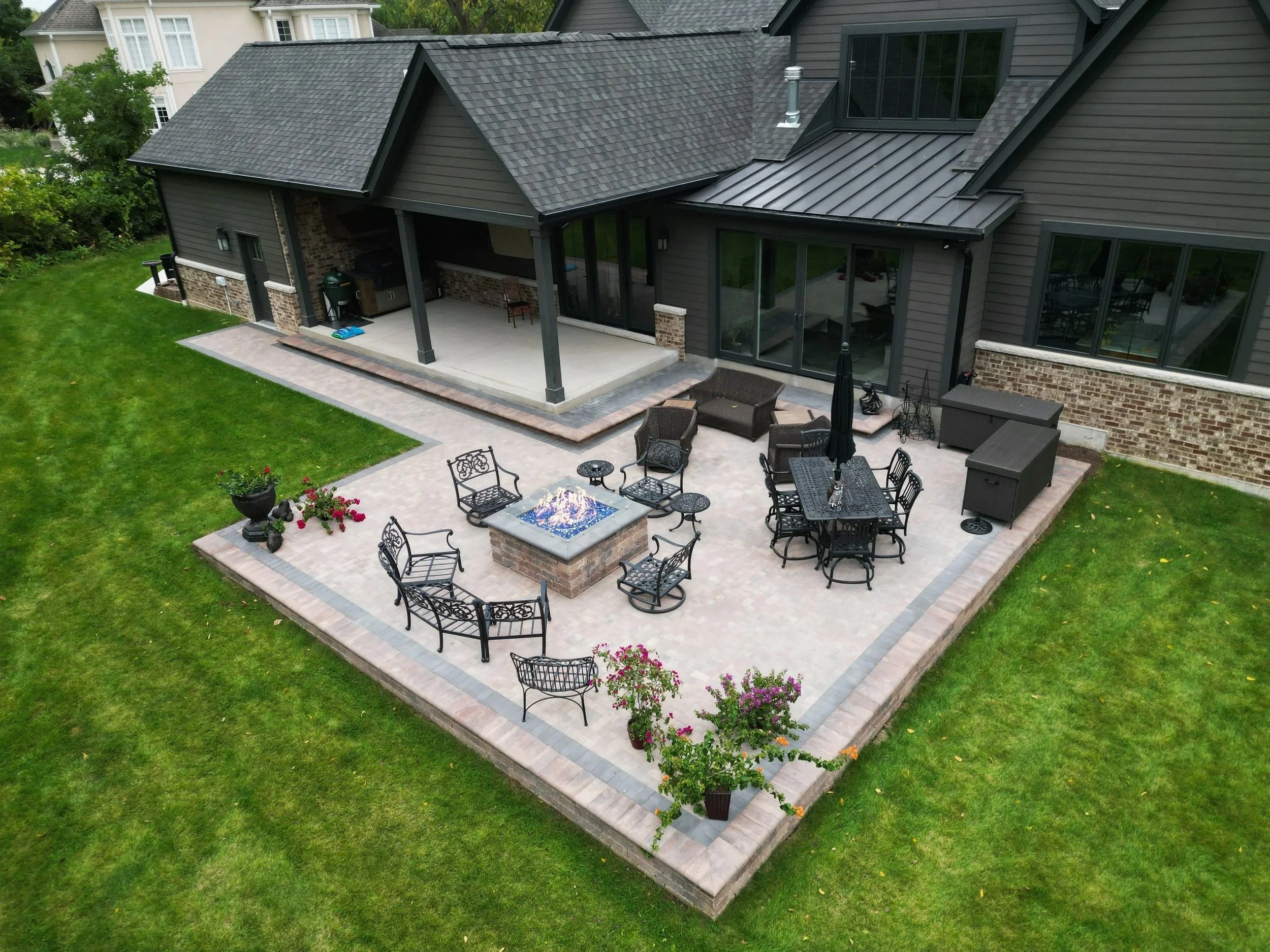 A backyard patio with outdoor furniture, fire pit, potted plants, and a covered porch area attached to a house with large windows.