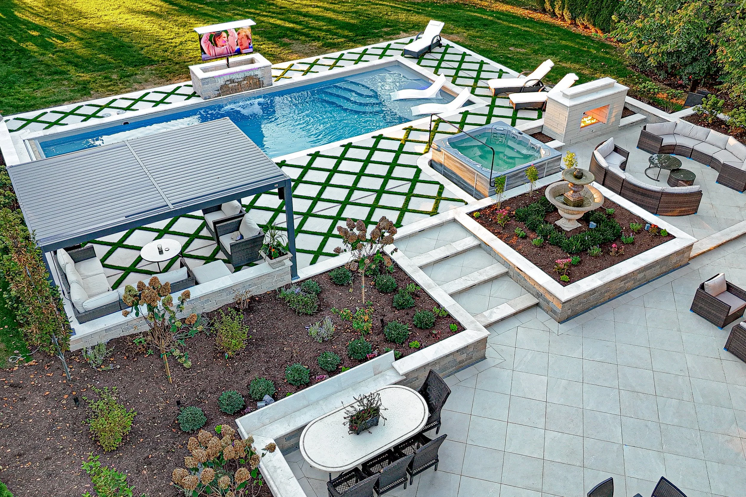 A backyard outdoor space with a swimming pool, hot tub, lounge chairs, a firepit, a covered seating area with a table, a patio with chairs, and landscaped garden beds, surrounded by a lawn and trees.
