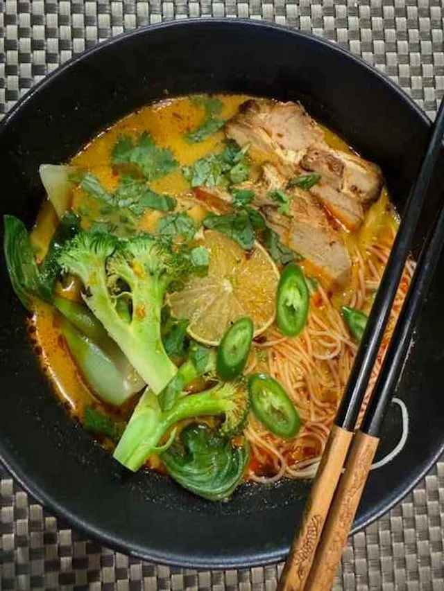 Spicy ramen with noodles, grilled pork, lime, bok choy, broccoli, and green chili slices in a black bowl with chopsticks