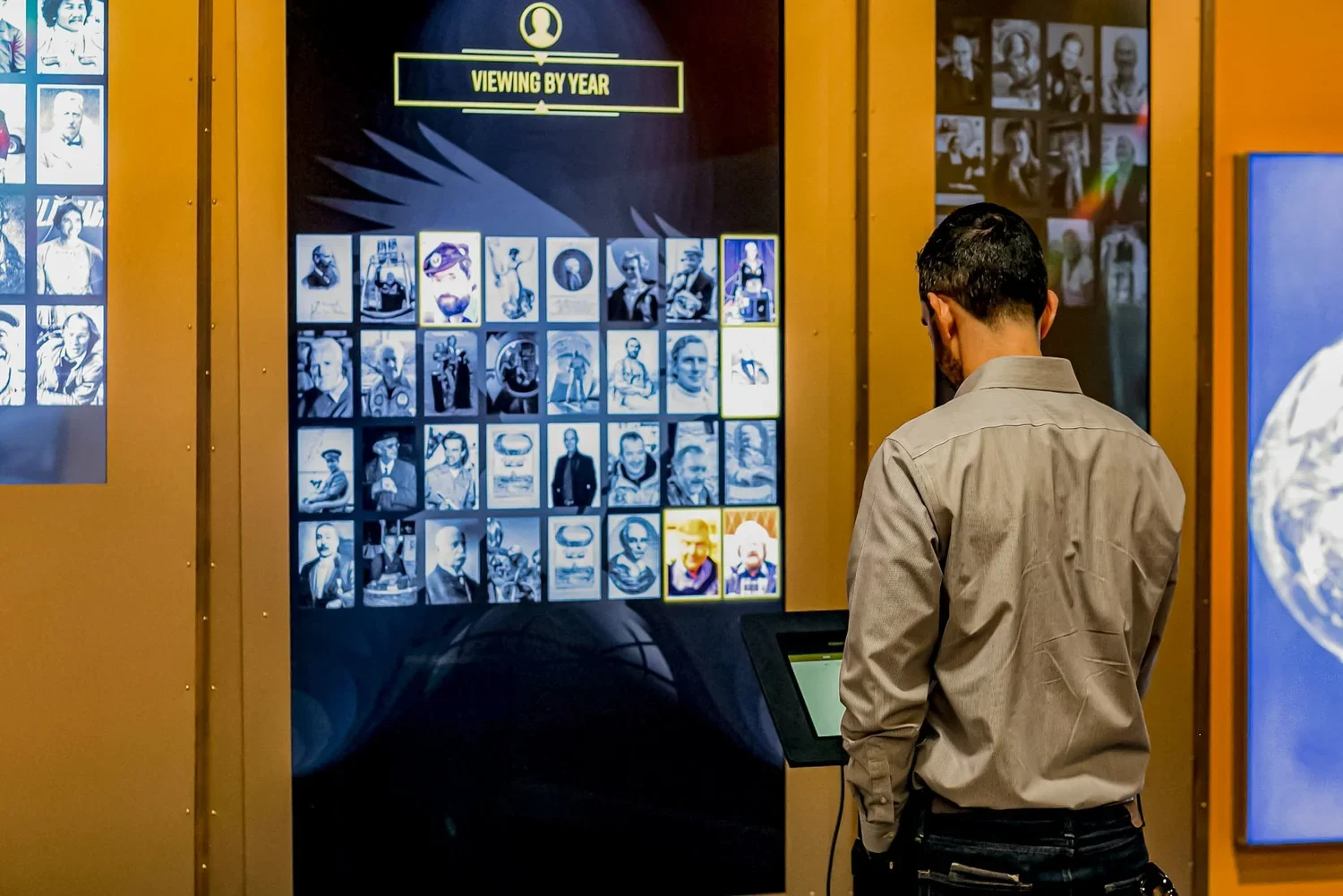 A man stands in front of a digital display of images organized by year, with a caption that reads 'Viewing by Year.' The display features multiple rows of black and white and color photographs of various people, and the man appears to be selecting or viewing the images.