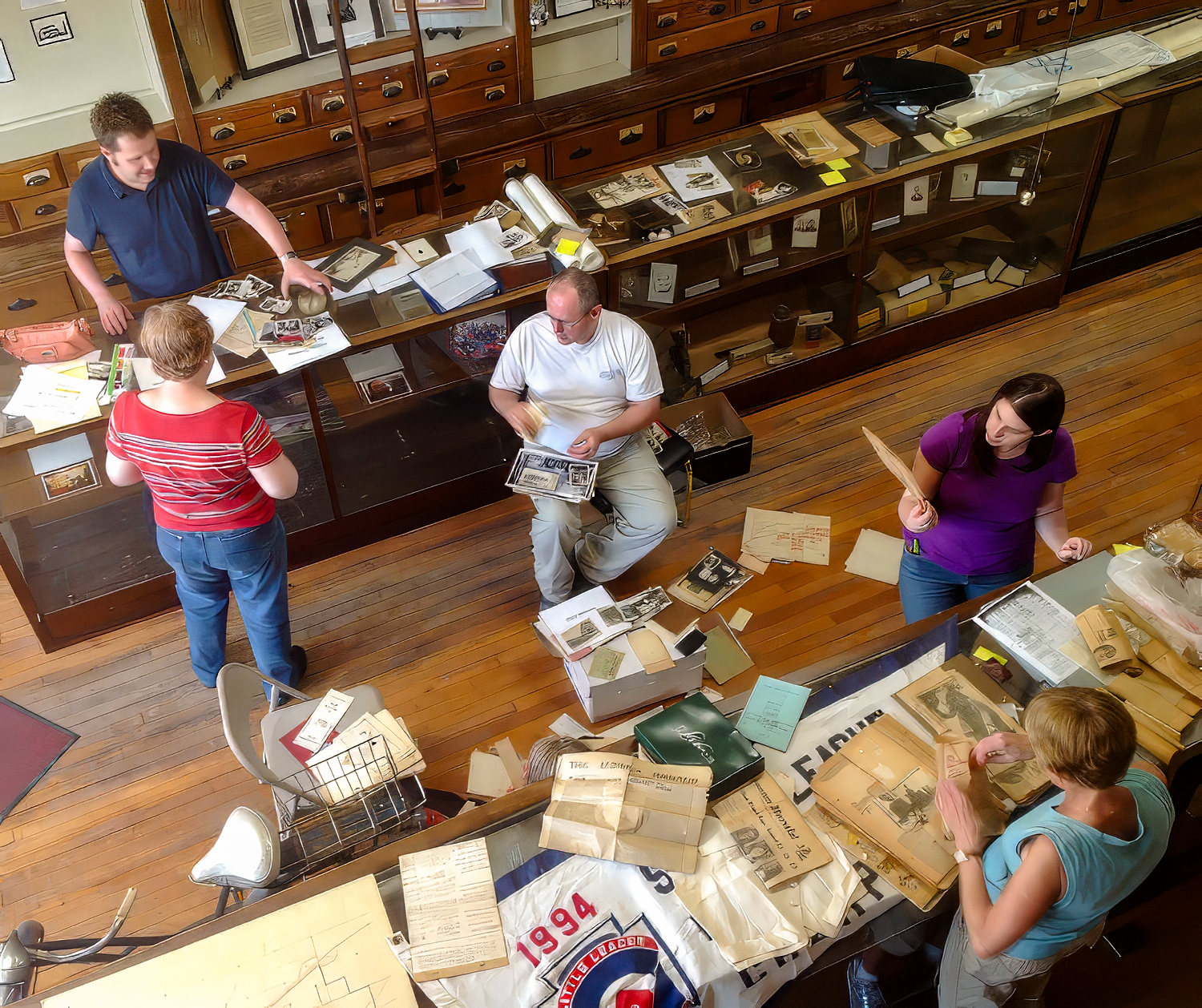 People sorting through cardboard boxes and old documents in a museum or archive room, surrounded by glass display cases filled with artifacts.