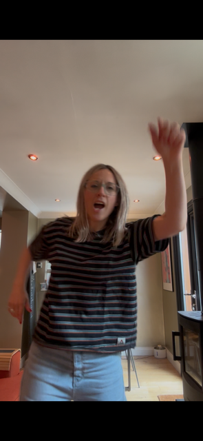 A young woman with long blonde hair, wearing a striped shirt and light-colored pants, appears to be smiling and raising her hand, possibly dancing or waving inside a living room.