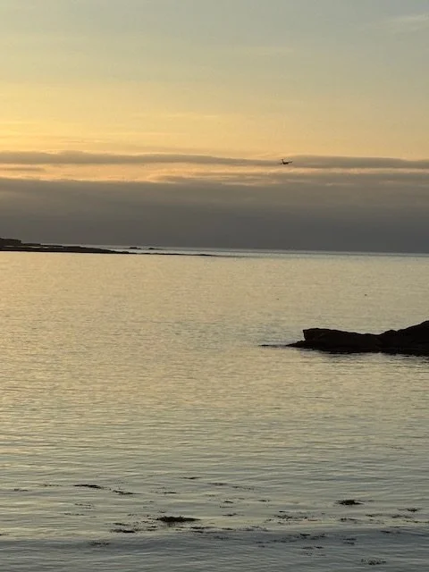 A serene beach scene at sunset with calm water, a rocky outcrop, and a small plane flying in the distance across the sky.