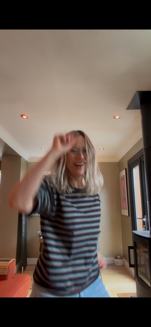 Woman with blonde hair wearing a striped shirt smiling and raising her hand in a living room.
