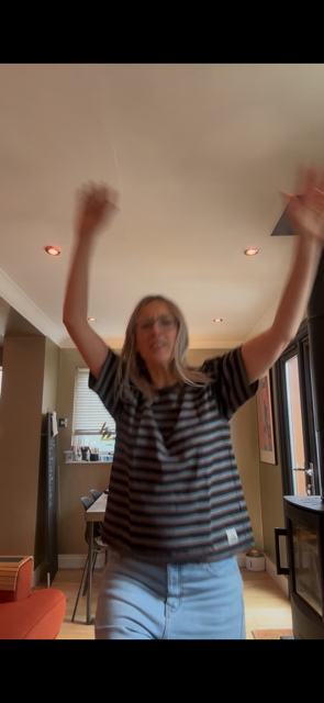 Woman standing indoors with arms raised, wearing a striped shirt and jeans.