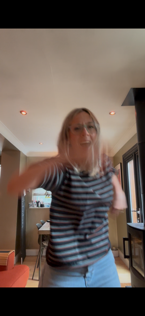 A woman with blonde hair and glasses, wearing a striped shirt, standing in a room with beige walls and large windows.