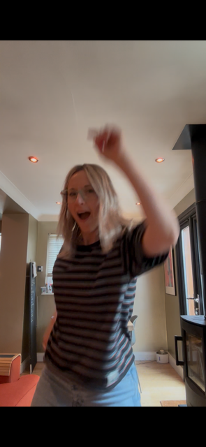 A woman with blonde hair wearing a striped shirt, smiling and raising her arm in a cheerful gesture inside a living room with modern decor.