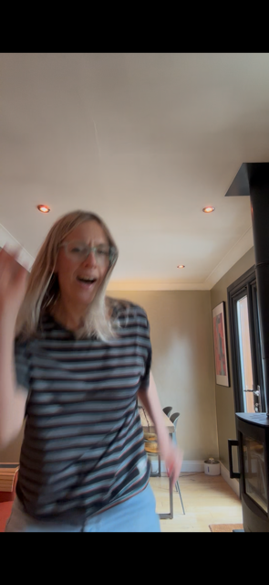 A woman with blonde hair and glasses, wearing a striped shirt, appears to be in motion inside a living room, smiling or laughing.