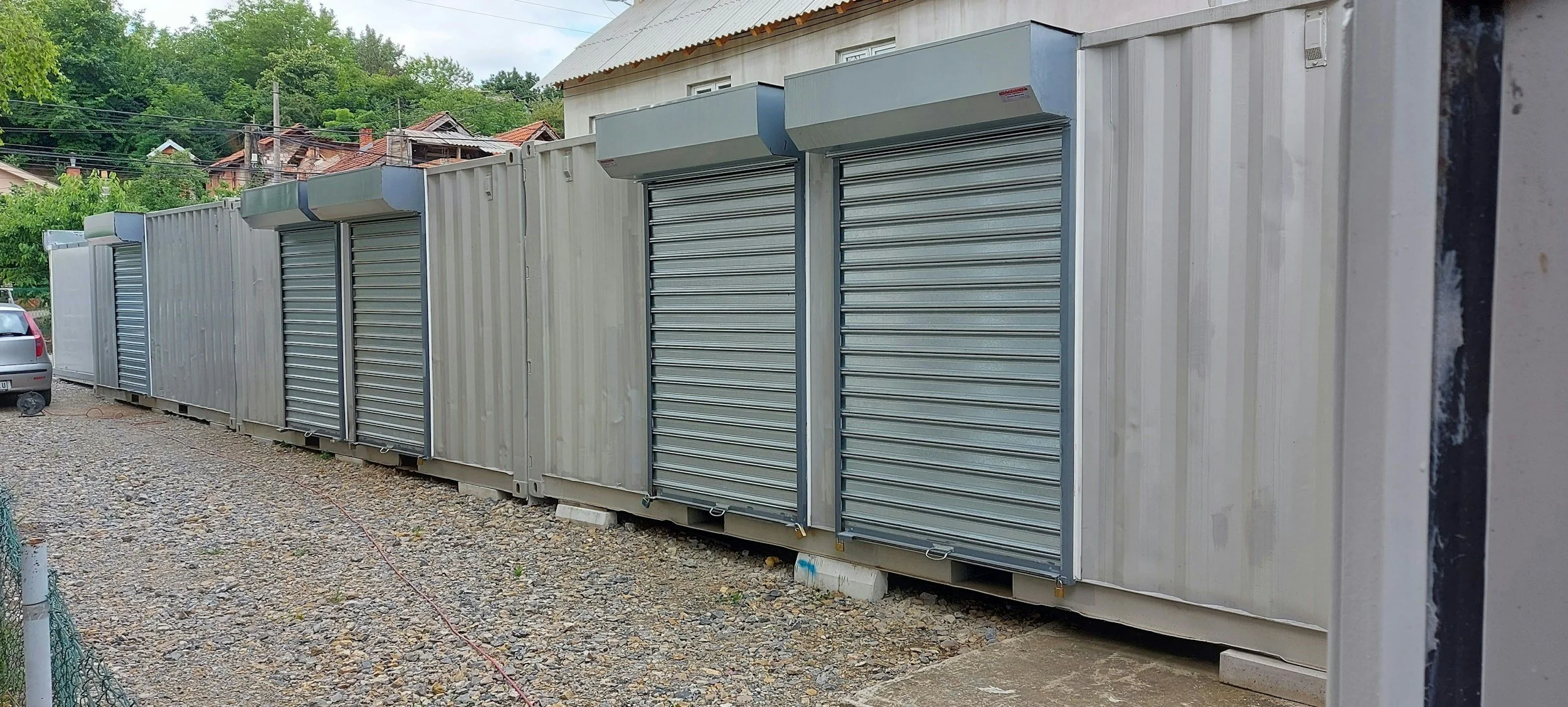 A row of metal storage units with roll-up doors and weather protection awnings, set on concrete blocks on a gravel surface with a car parked nearby.
