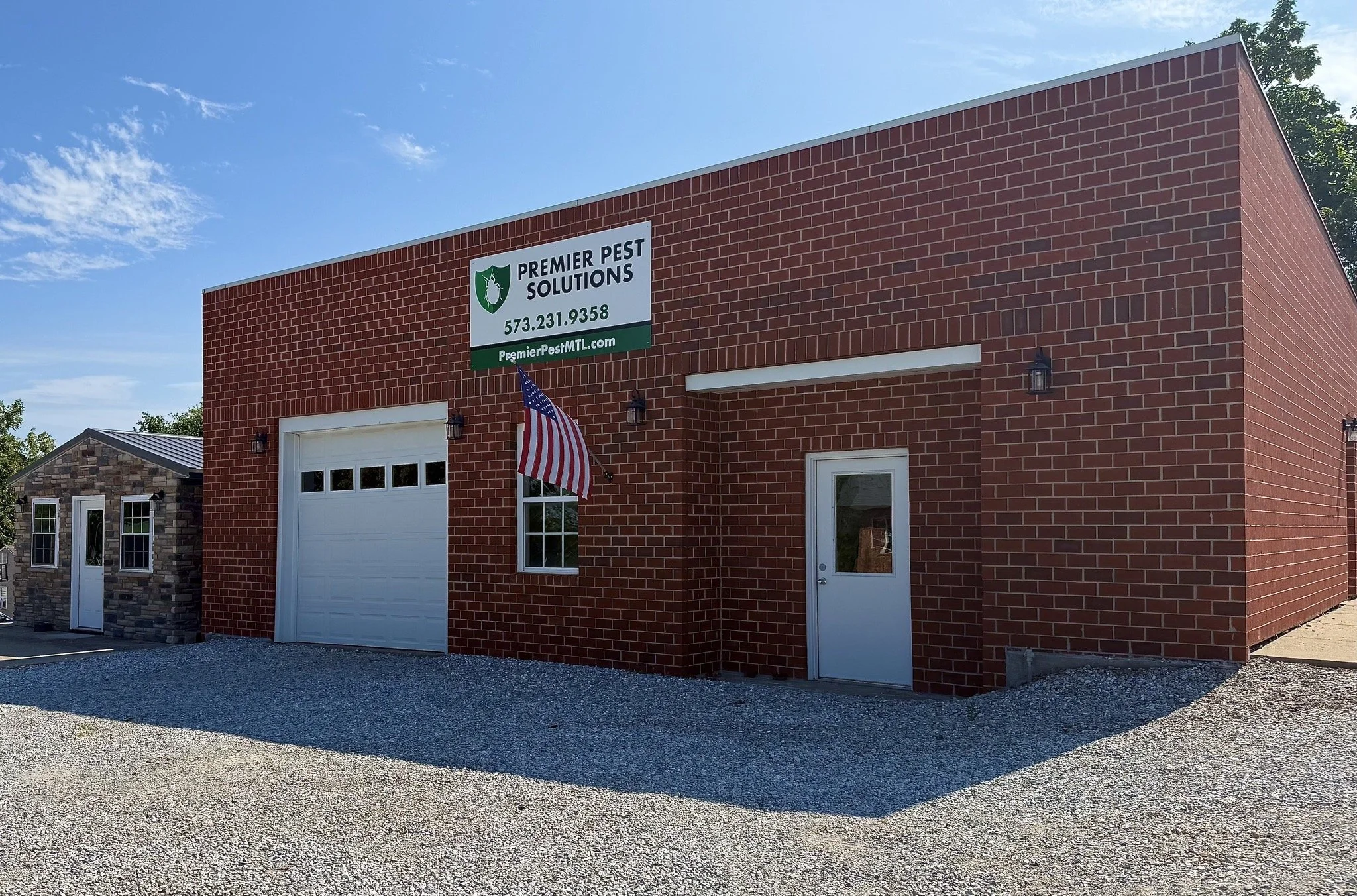 A red brick building with two garage doors, two windows, and two exterior lights, with an American flag hanging near one of the doors and a sign that reads 'Premier Pest Solutions' with contact information. The sky is clear and blue with a few clouds.