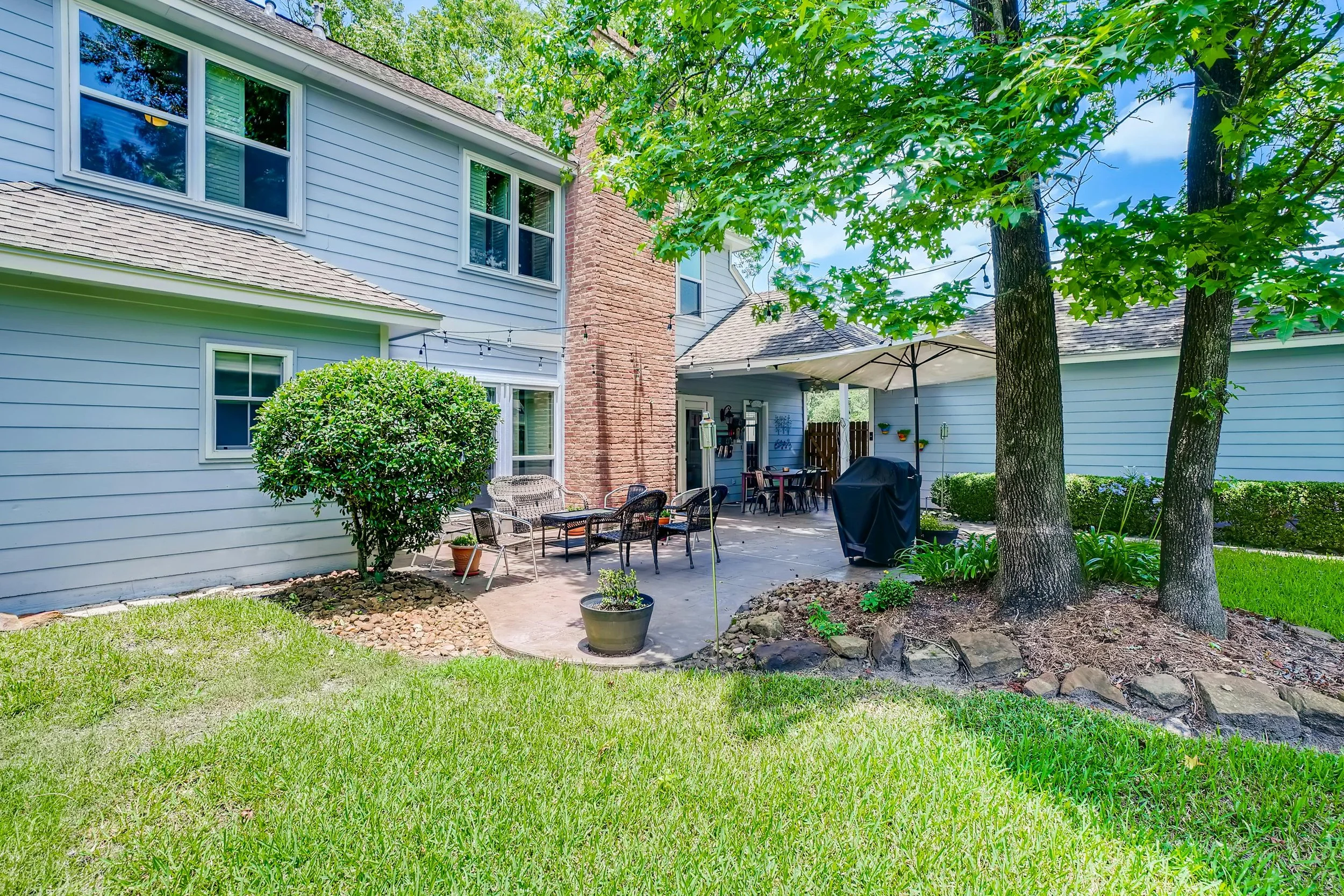 Backyard patio area with outdoor furniture, umbrella, and potted plants, surrounded by green trees and a blue house with white trim. Protect lawns like this with Premier Pest Solutions