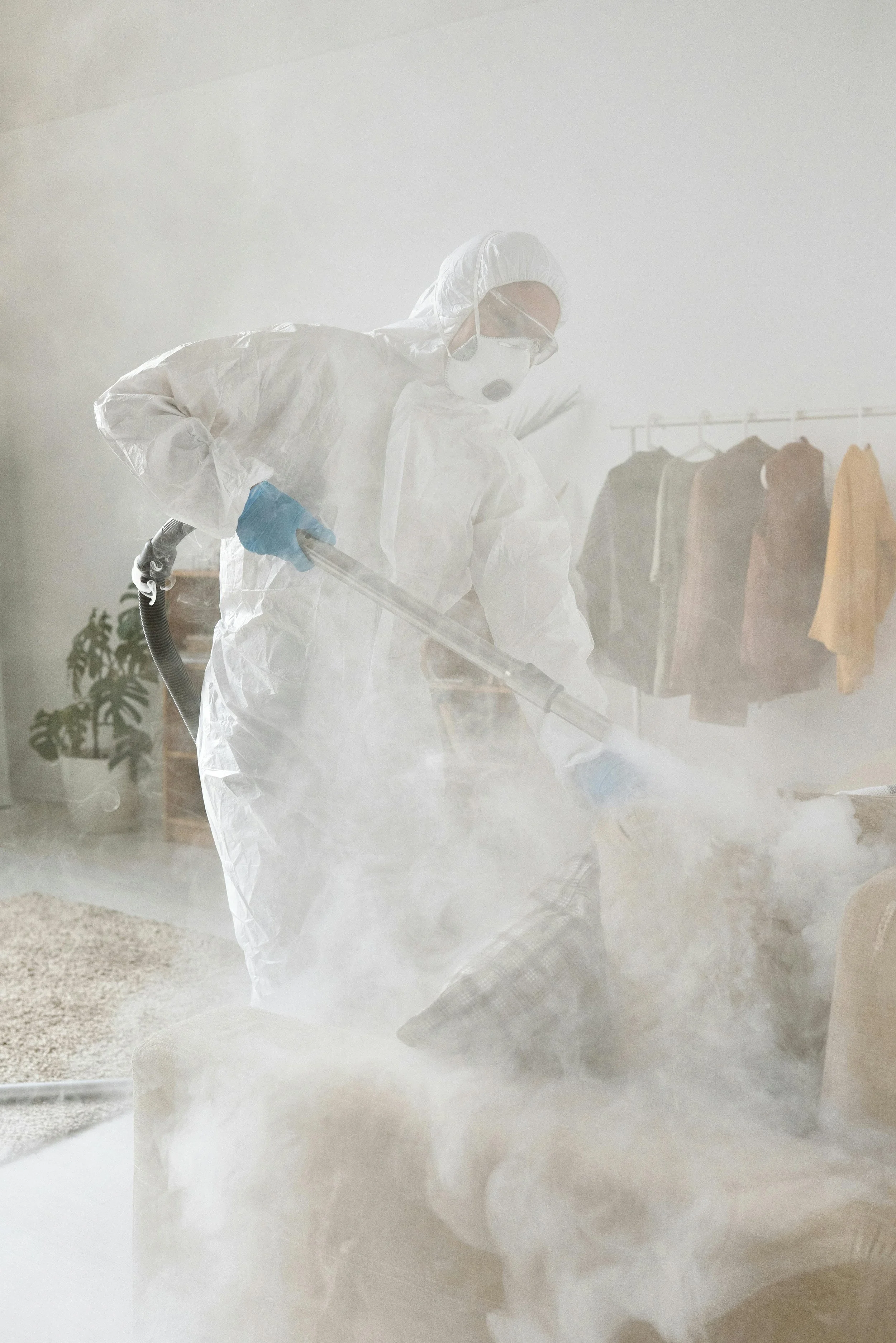 Person in protective suit using fogging equipment in a room filled with fog or mist, with a clothing rack and plants in the background.