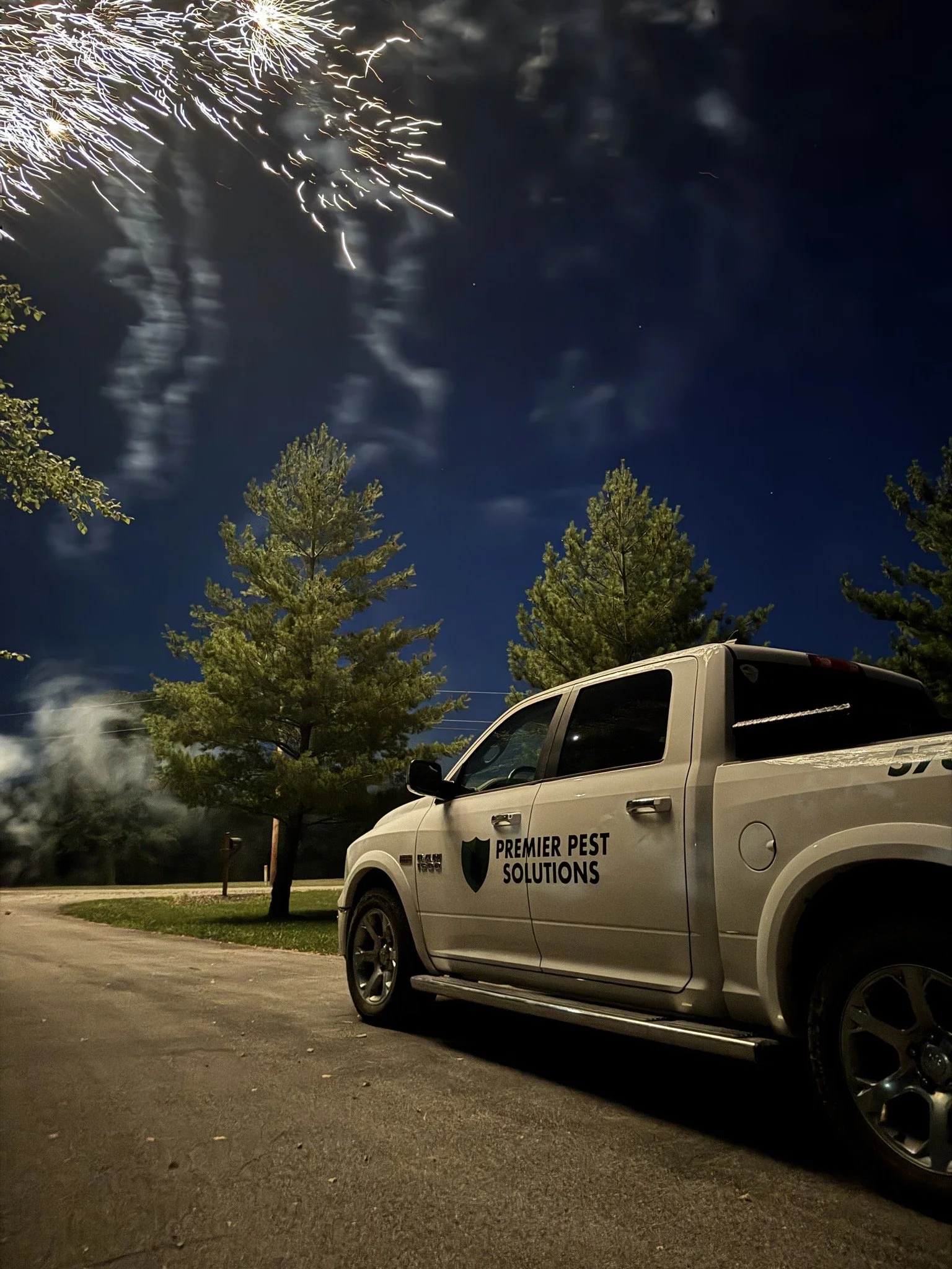 Nighttime scene showing fireworks in the sky, with smoke and clouds. In the foreground, there is a silver pickup truck with the logo and text 'PREMIER PEST SOLUTIONS' on the door, parked on a paved surface next to some green trees.