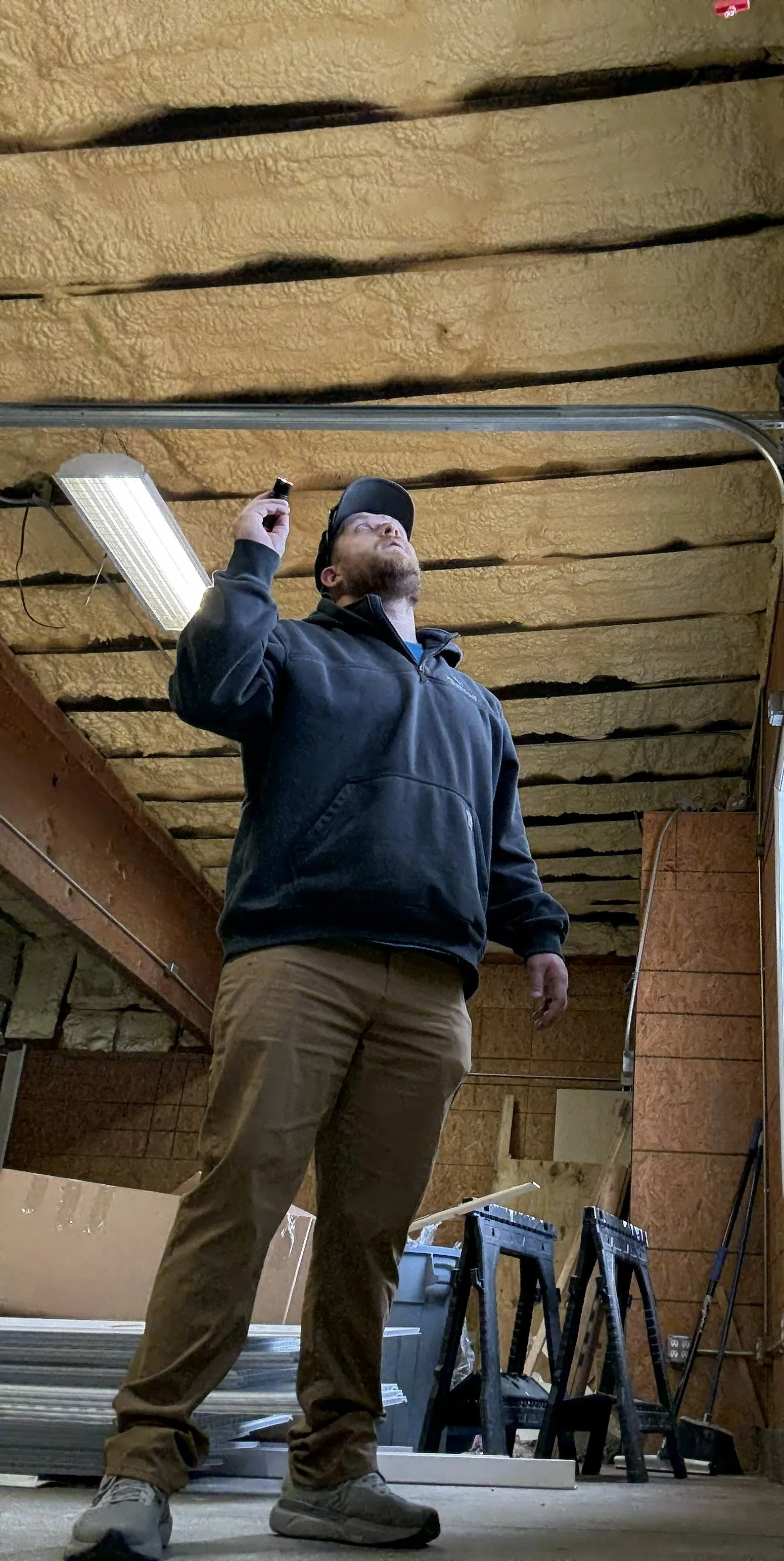 Man in black hoodie and tan pants inspecting insulation on ceiling in an attic.