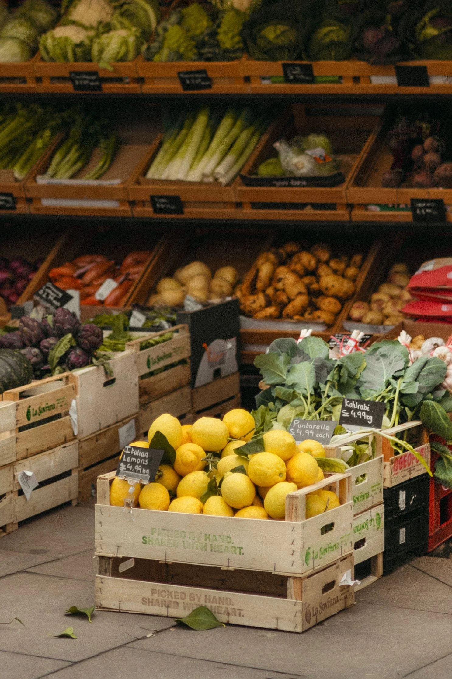 A display of fresh fruits and vegetables at a market, including a wooden crate of yellow lemons labeled as Italian Lemons, with other greens and produce behind them.