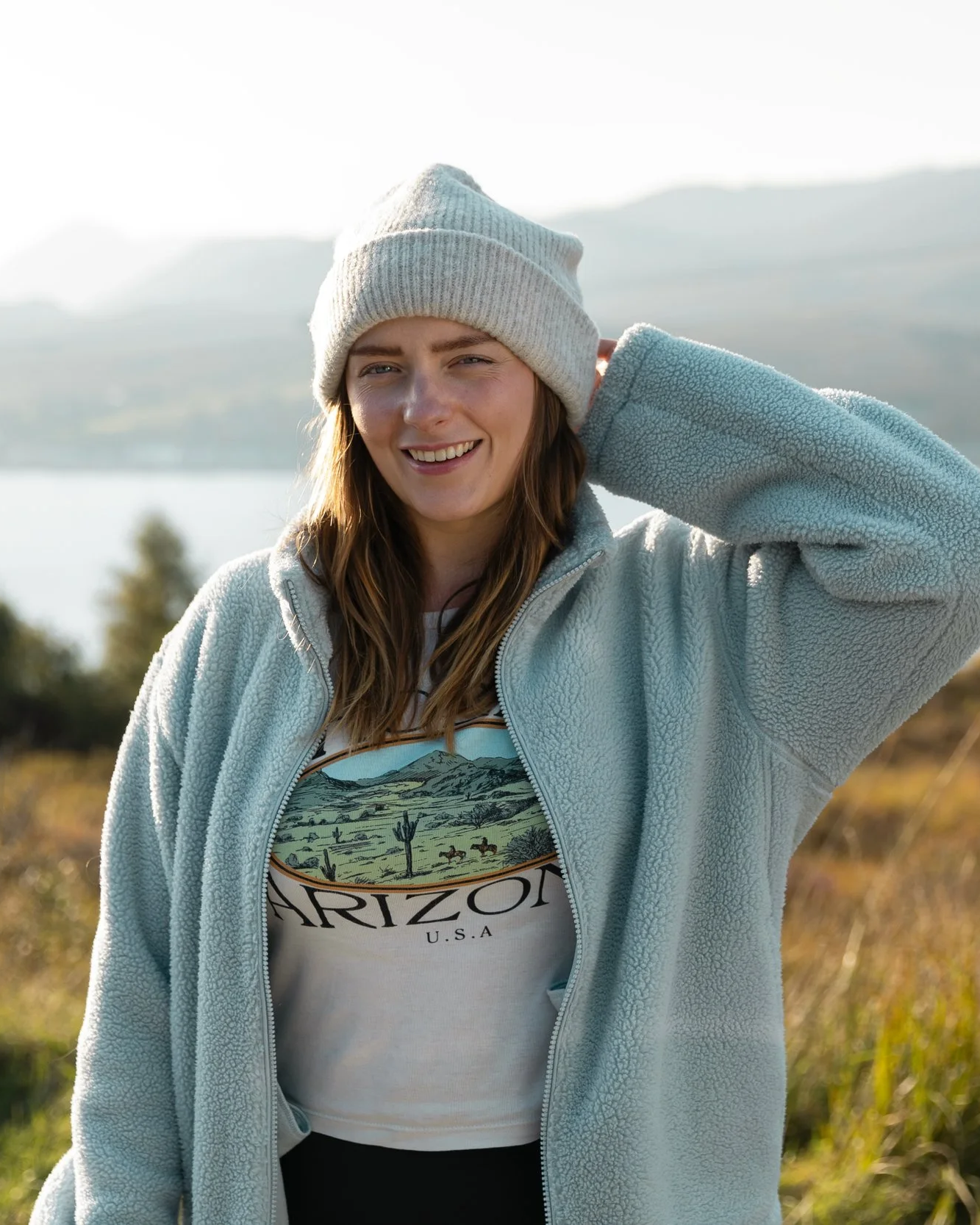 Young woman smiling outdoors, wearing a beanie, fleece jacket, and a t-shirt with 'Arizona U.S.A.' text, standing in a scenic landscape with water, trees, and mountains in the background.